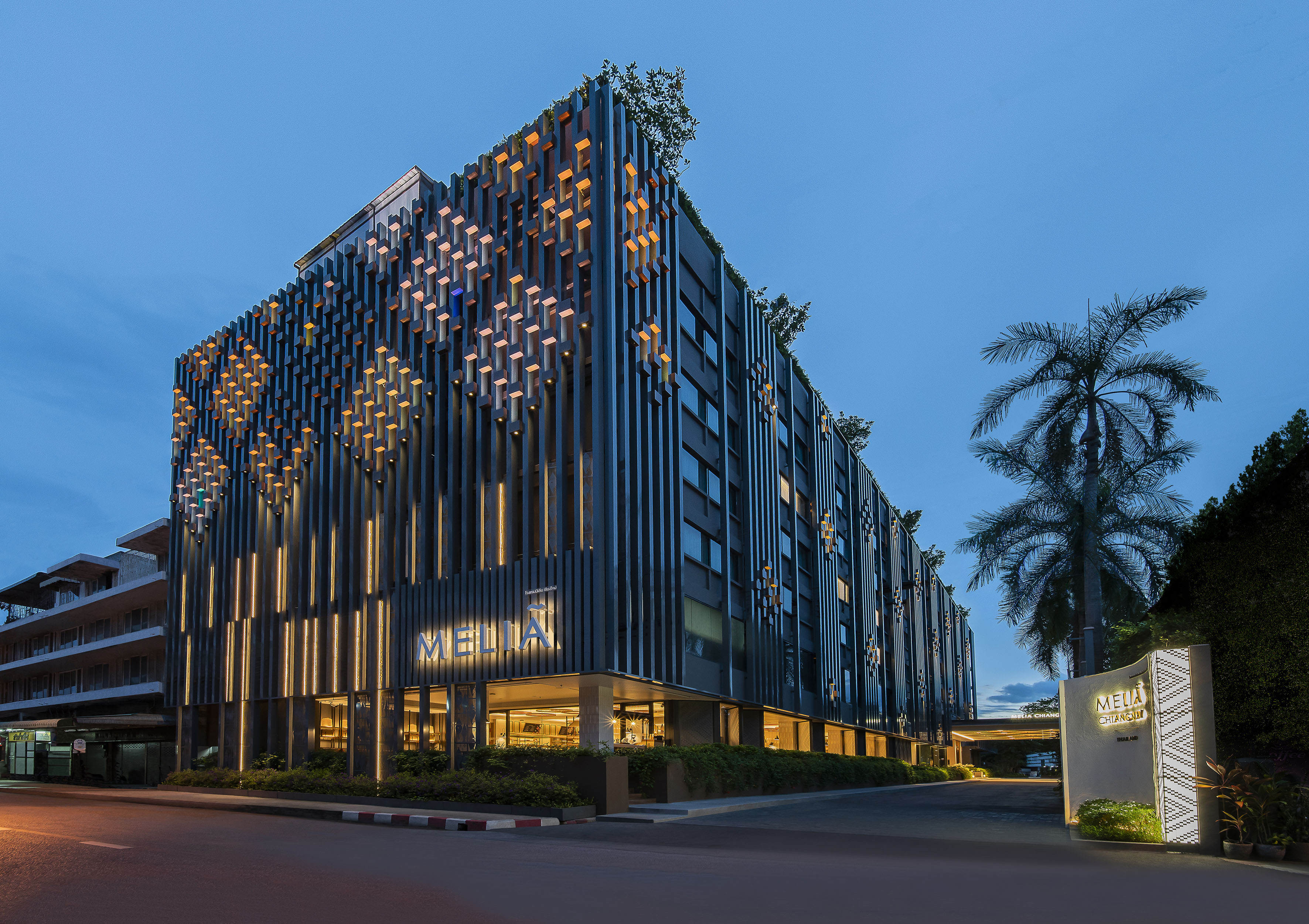 Modern Meliá Chiang Mai hotel at dusk, illuminated facade with vertical accents and palm tree.