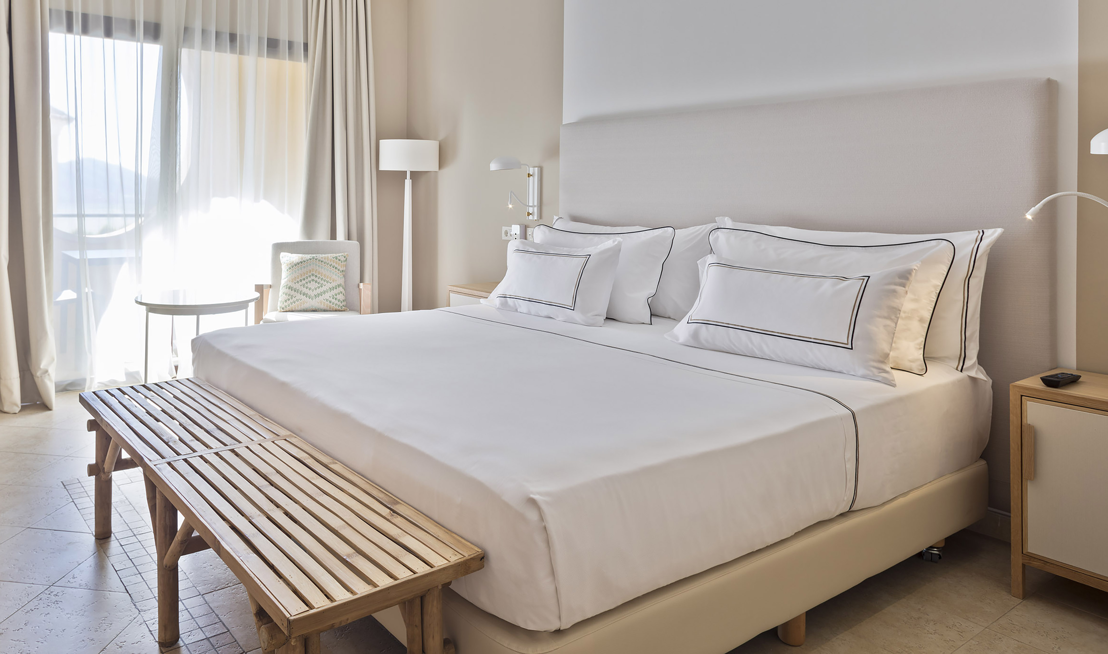 Luxury hotel bedroom with white linens, wooden bench, and bright window.