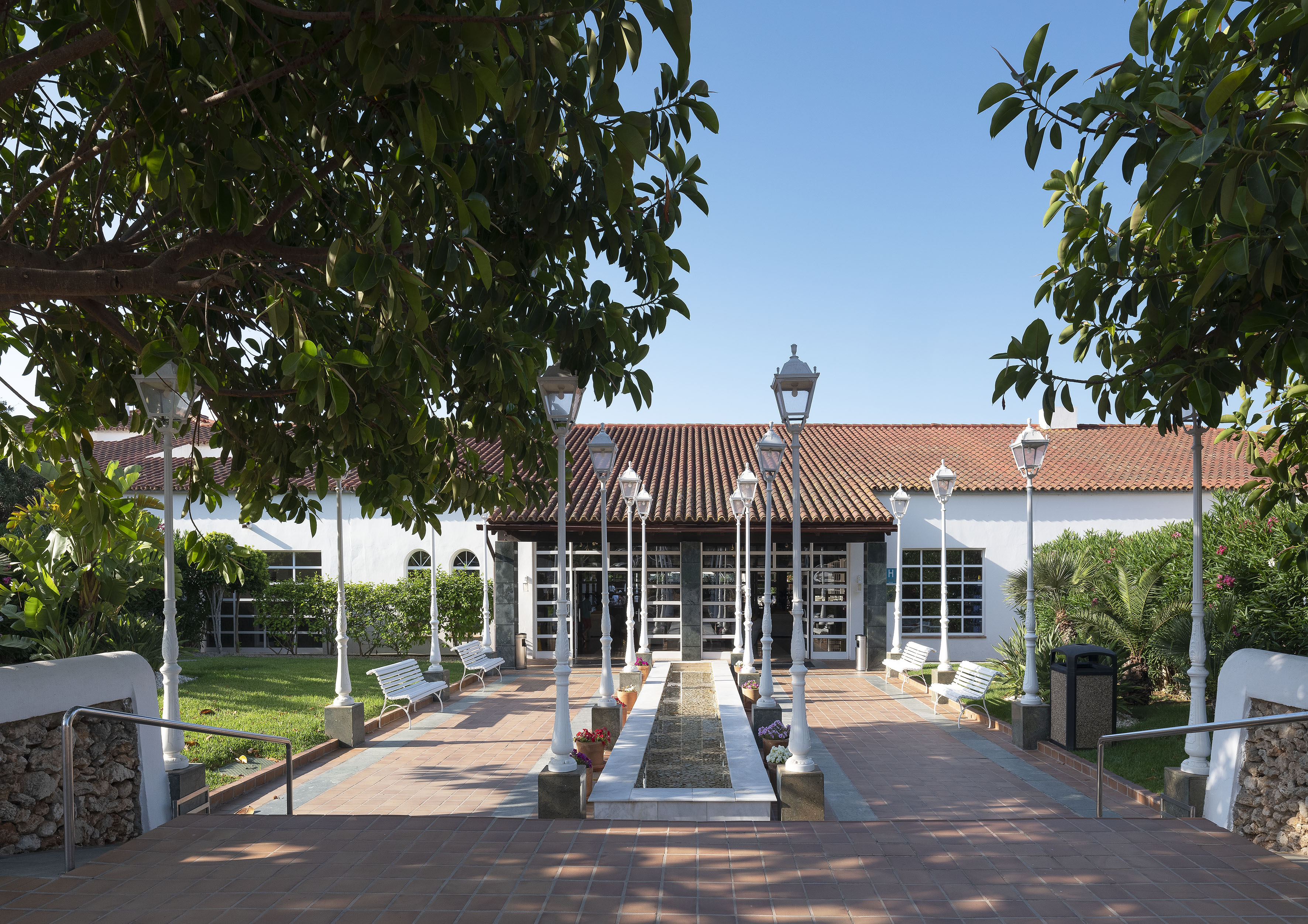 Sunny resort entrance with white building, red tile roof, central fountain, and lampposts.