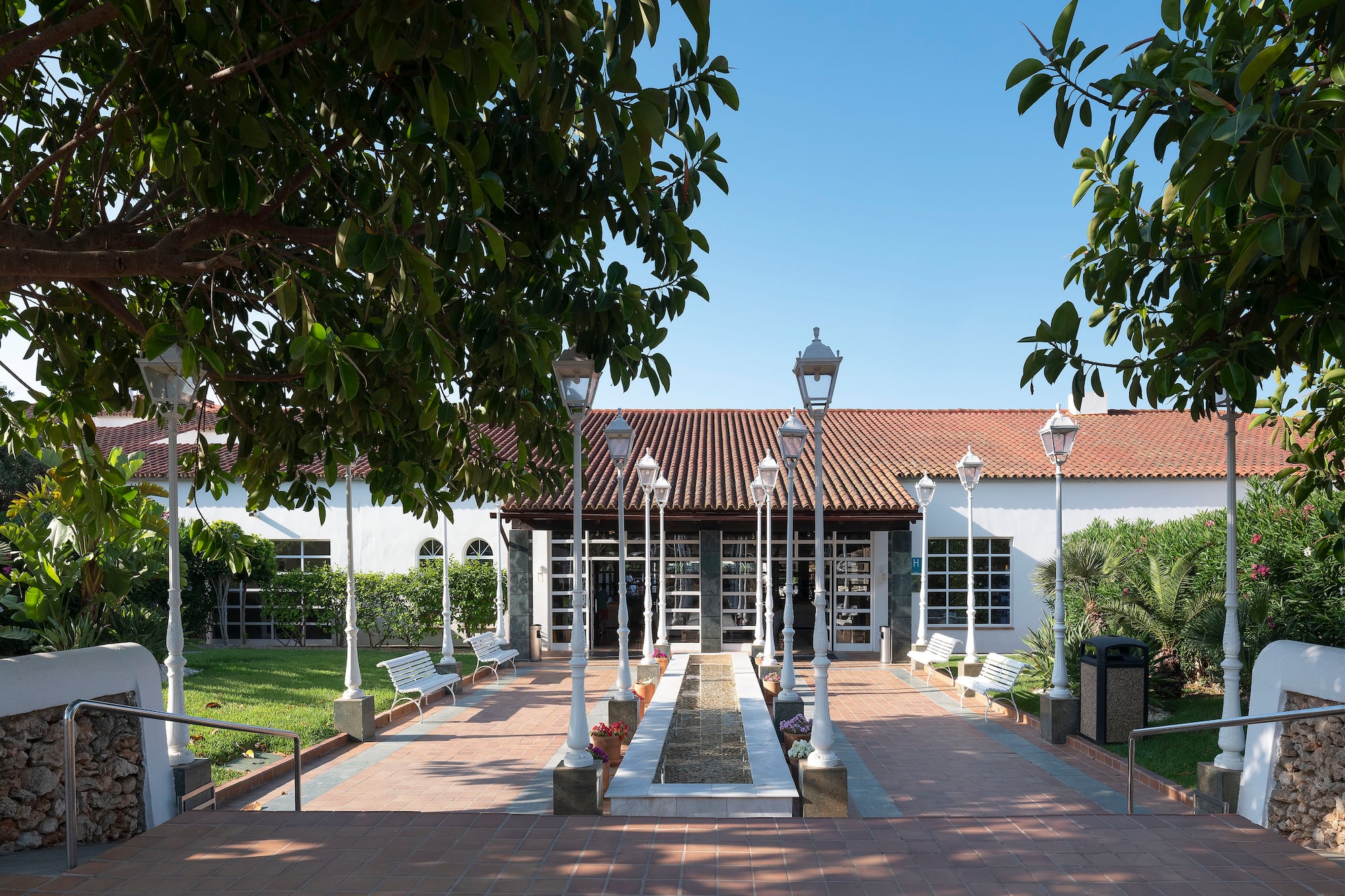 Sunny resort entrance with white building, red tile roof, central fountain, and lampposts.