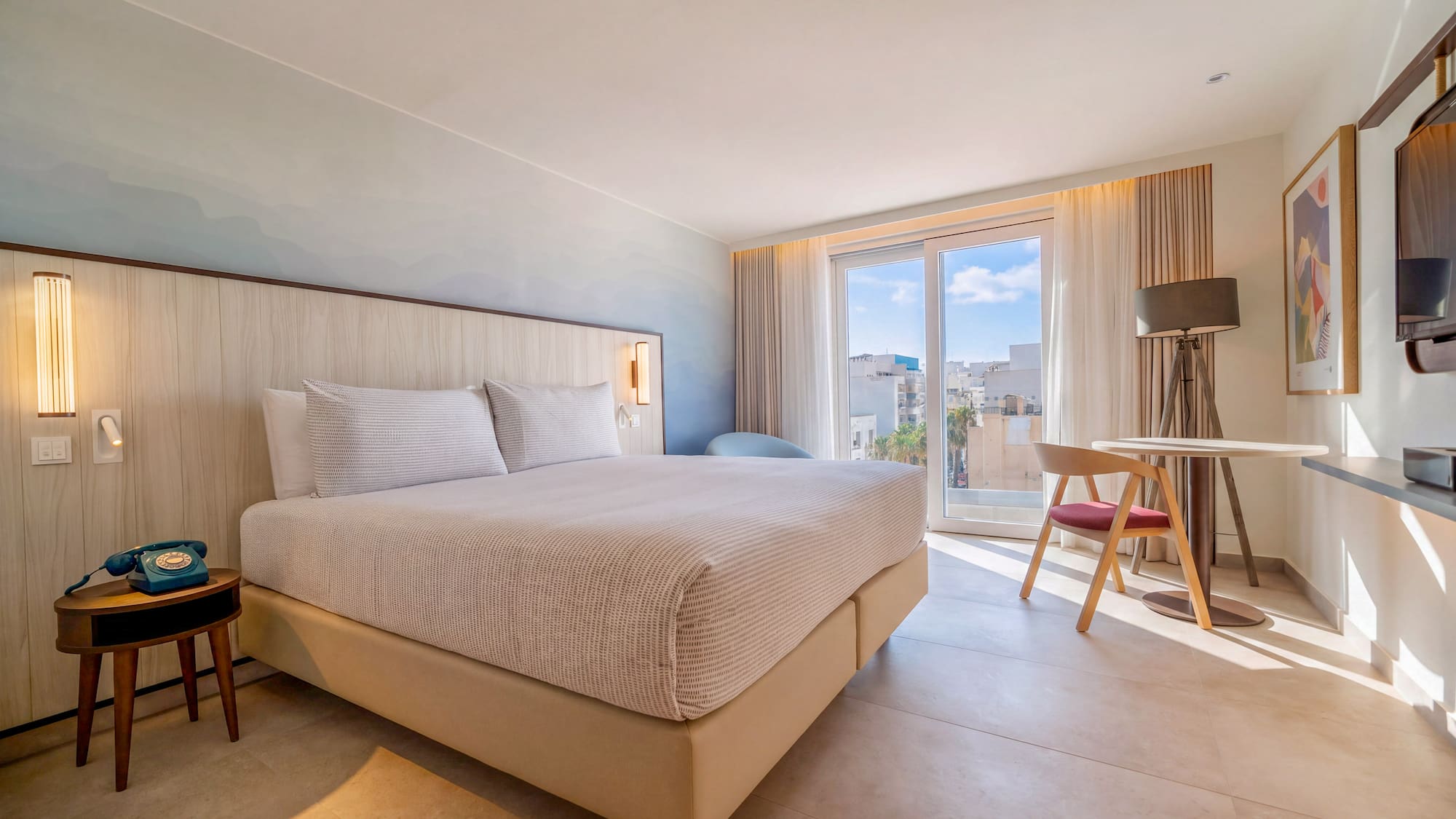 Bright modern hotel room, king bed, blue wall, balcony city view.