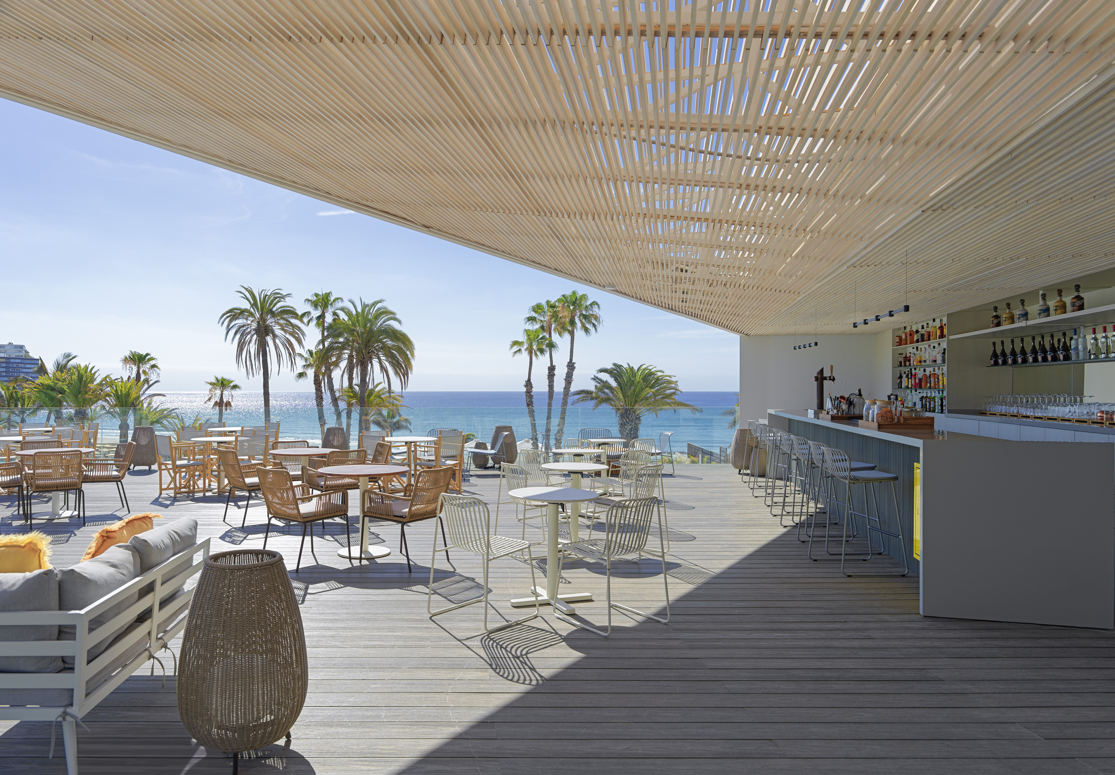 Sunny beachfront terrace with outdoor bar, lounge seating, ocean view, palm trees.