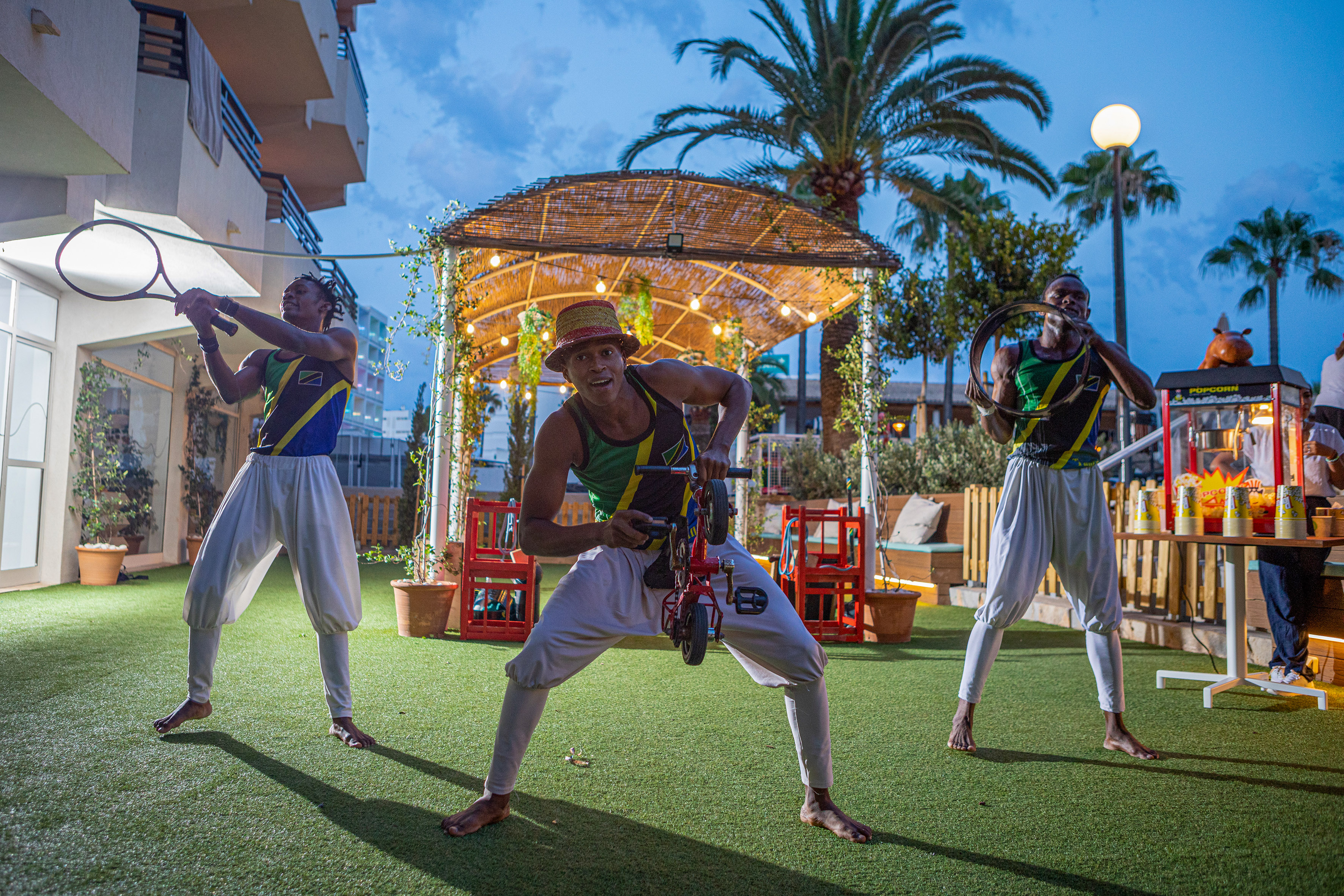 Performers with tennis racket, mini bike, hoop at an outdoor evening show.