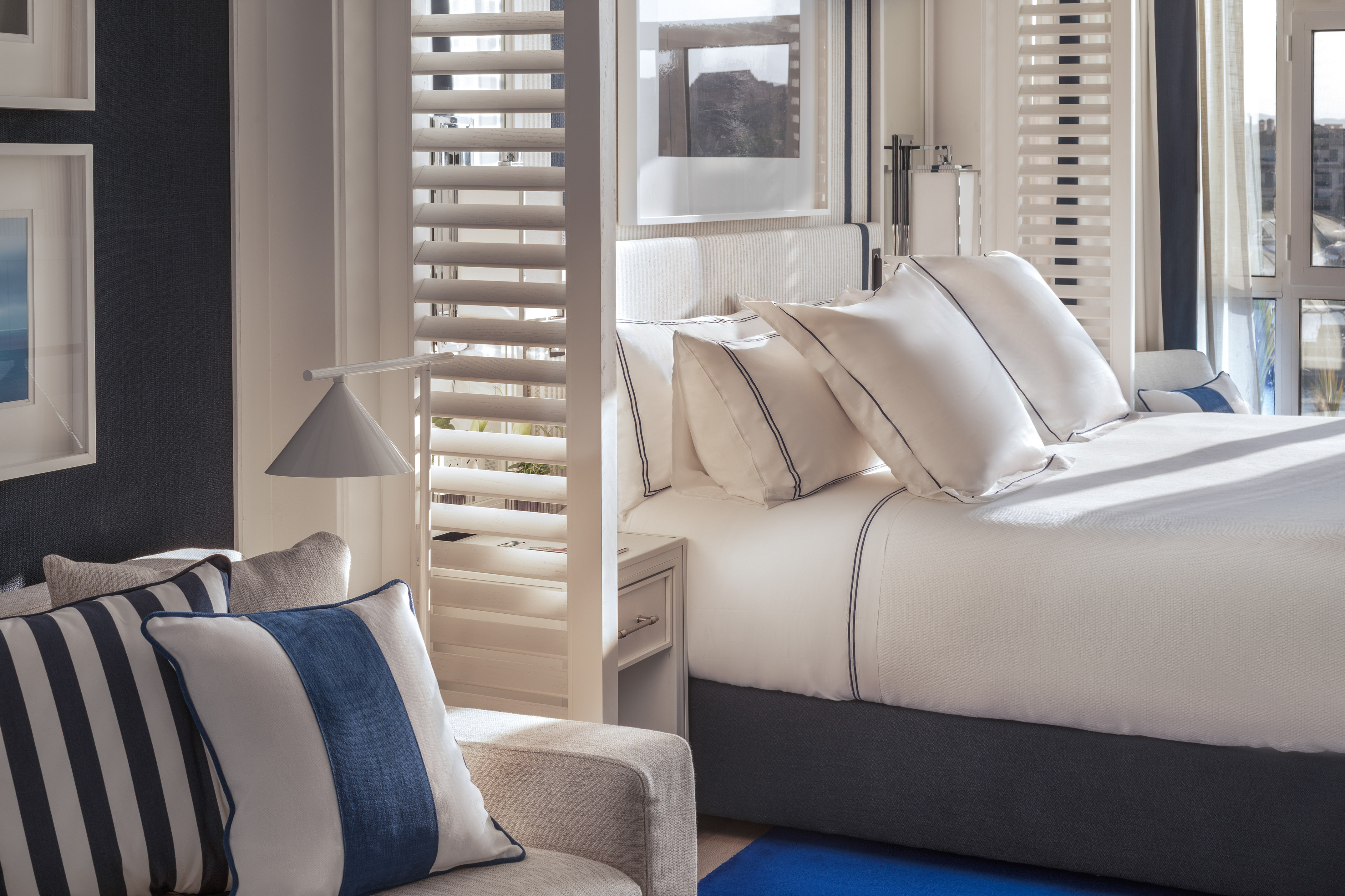 Coastal bedroom with white bed, blue striped pillows, and bright plantation shutters.