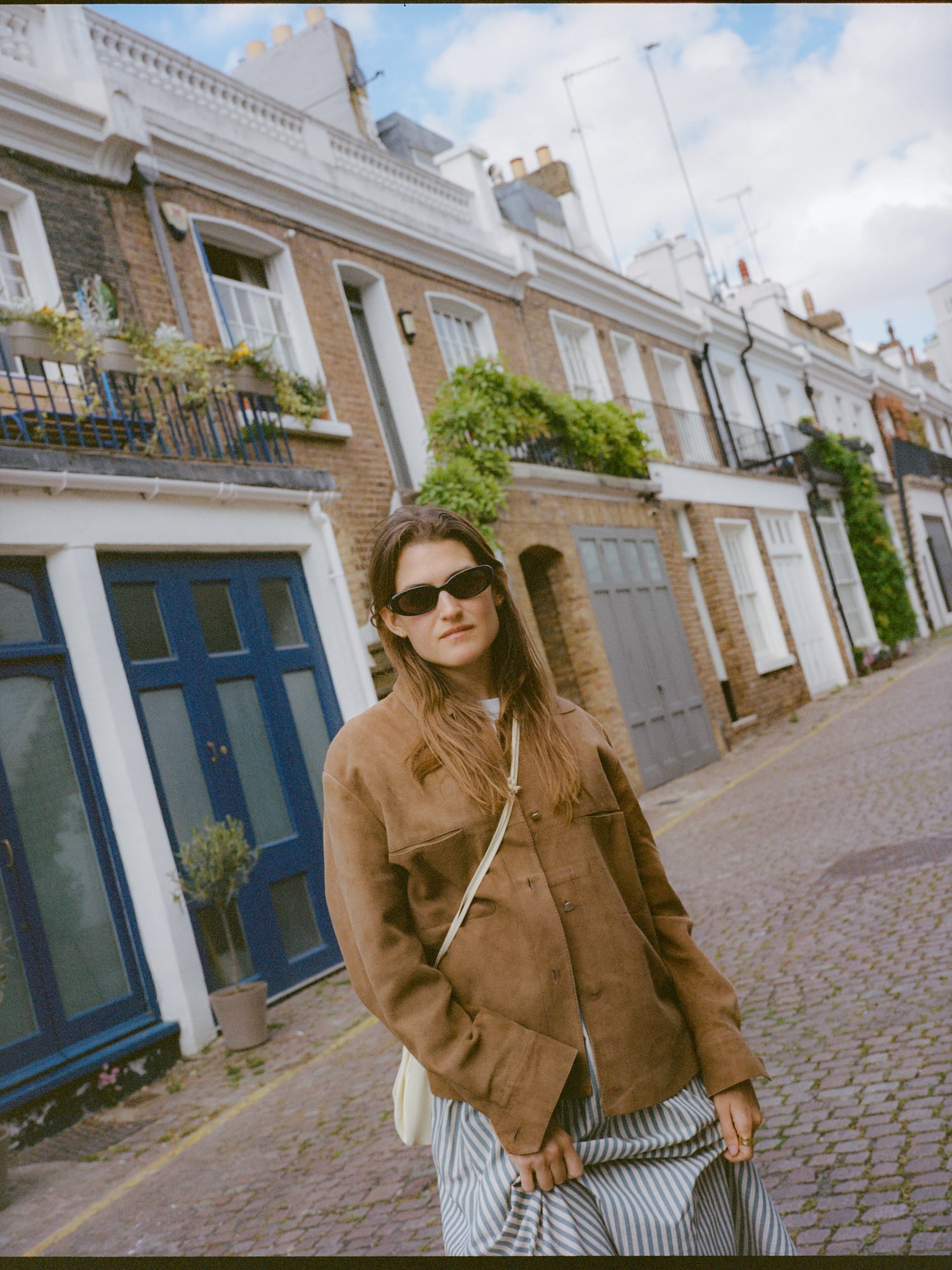 Woman in brown jacket and sunglasses on cobblestone street, brick houses.