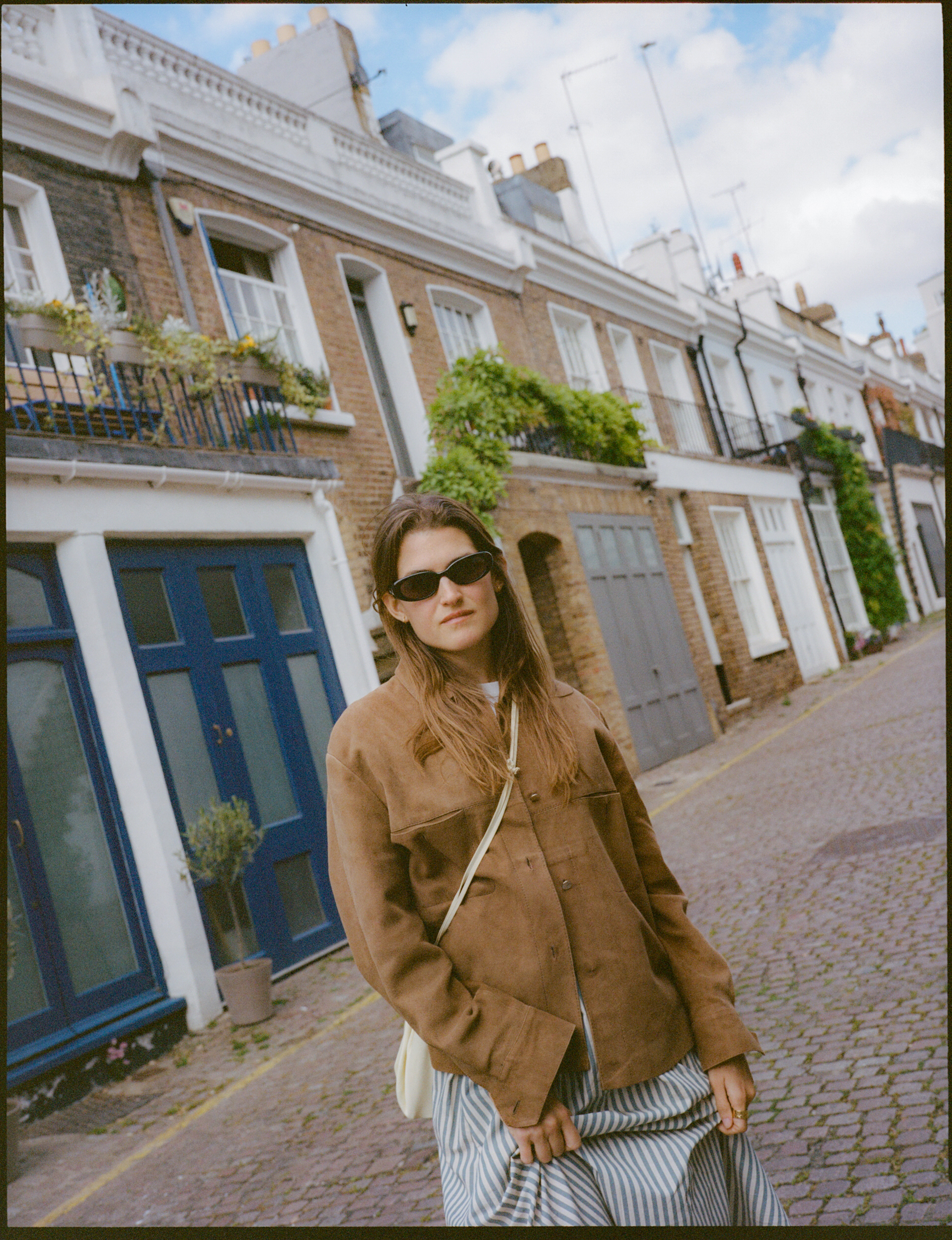 Woman in brown jacket and sunglasses on cobblestone street, brick houses.