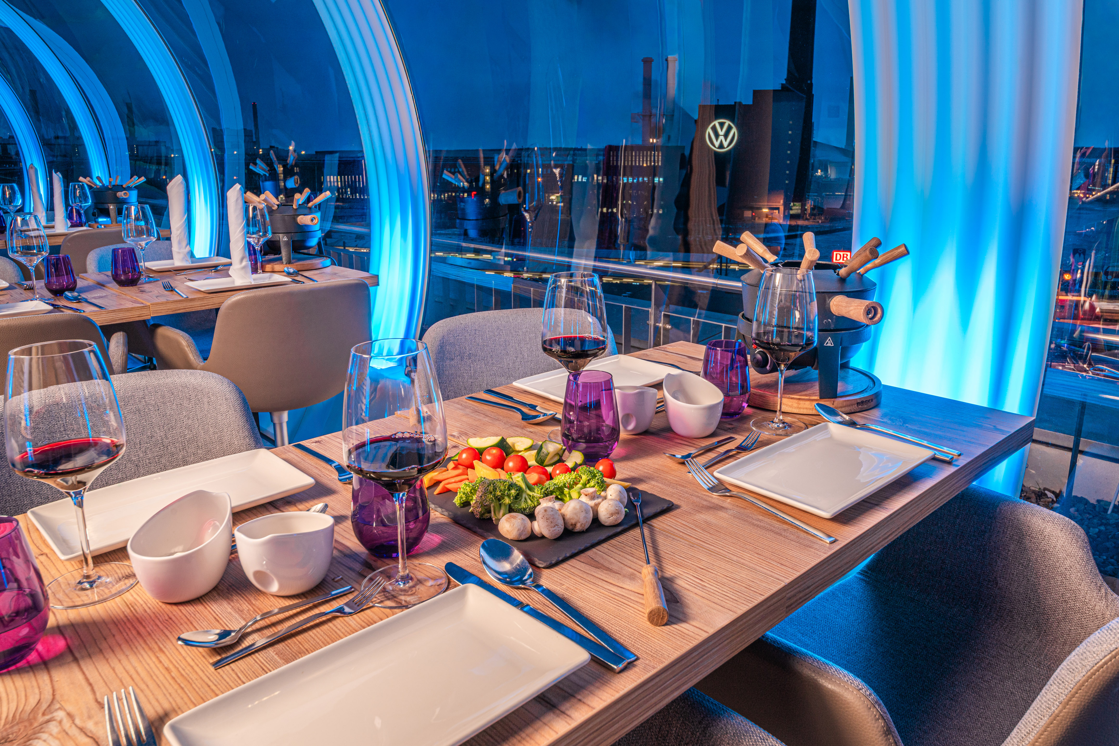 Modern restaurant table with fondue, fresh vegetables, wine, and city view.