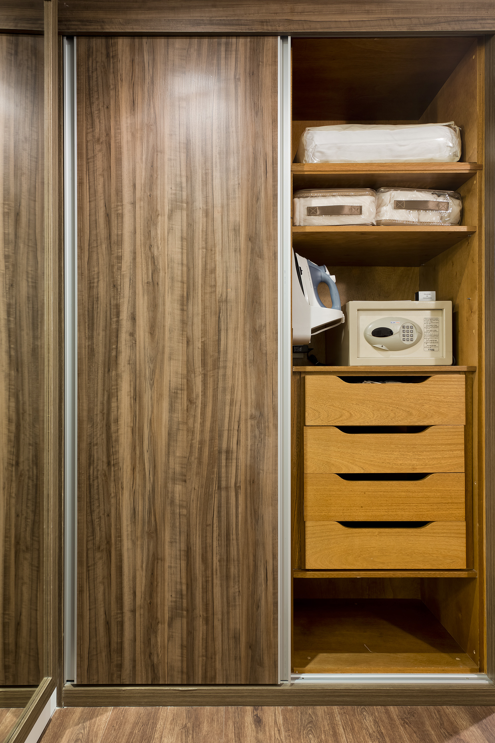 Open wooden closet with safe, iron, drawers, and folded bedding.