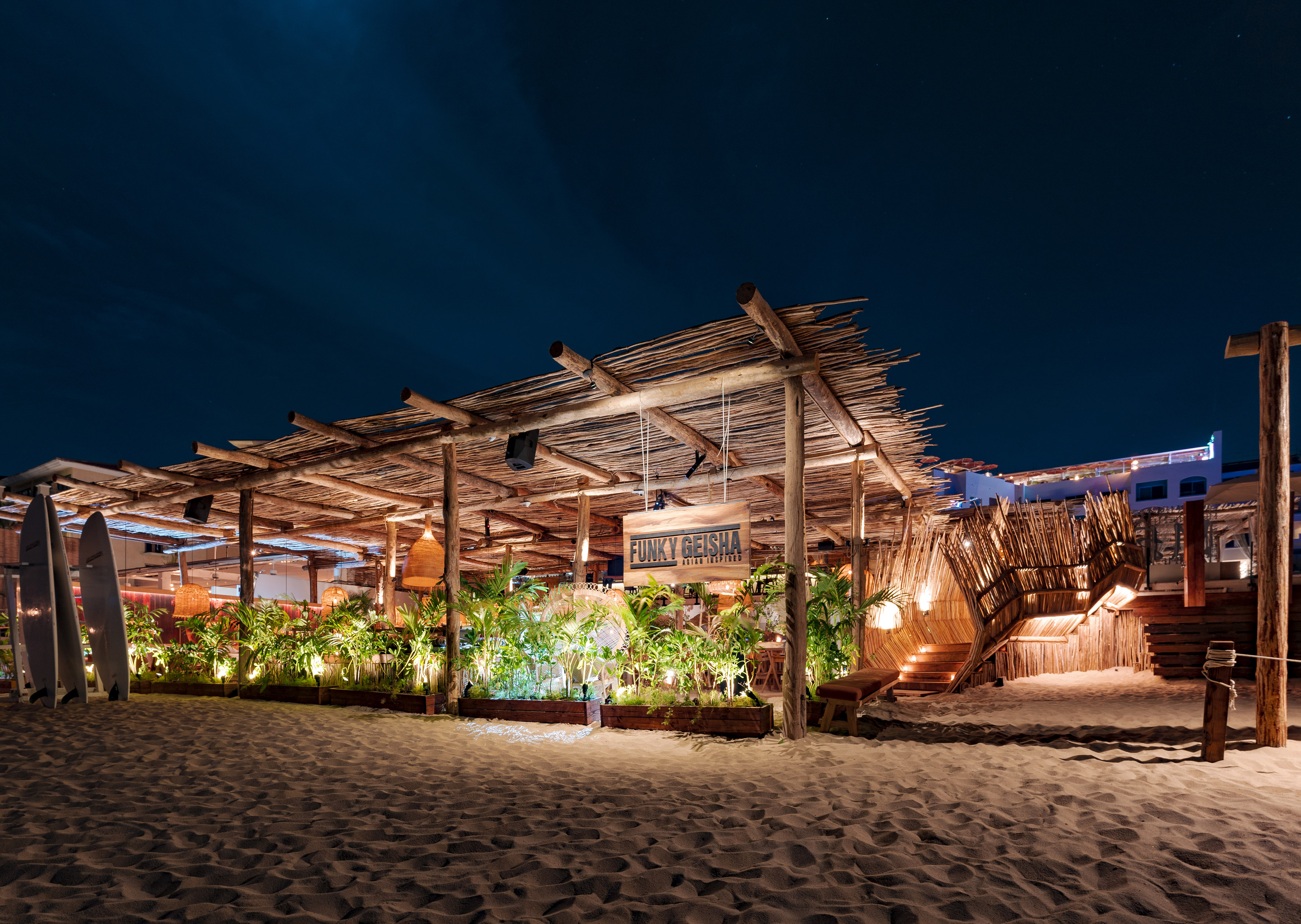 Tulum-style Funky Geisha beach restaurant at night, thatched roof, sandy beach.