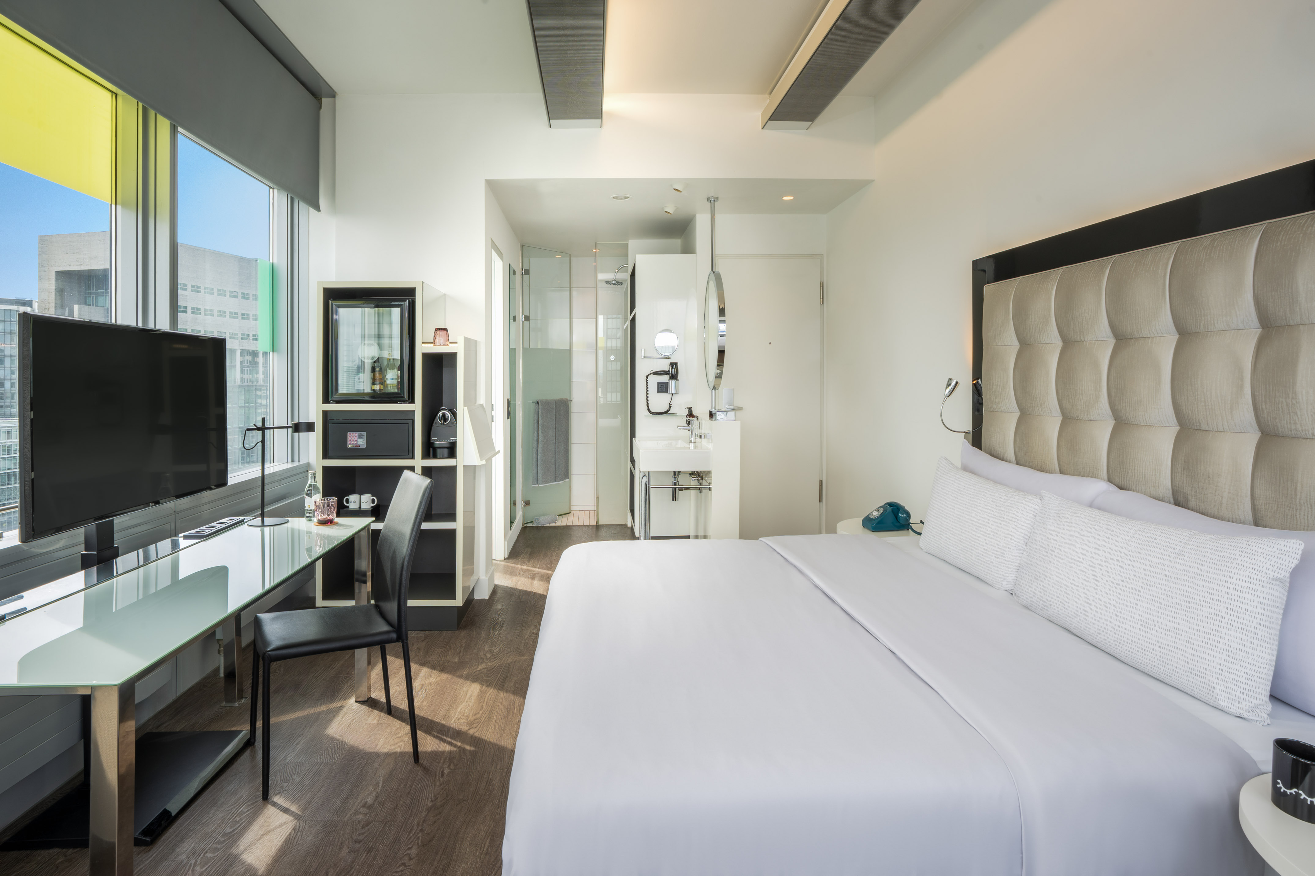 Modern hotel room with bed, glass desk, TV, city view, and integrated shower.