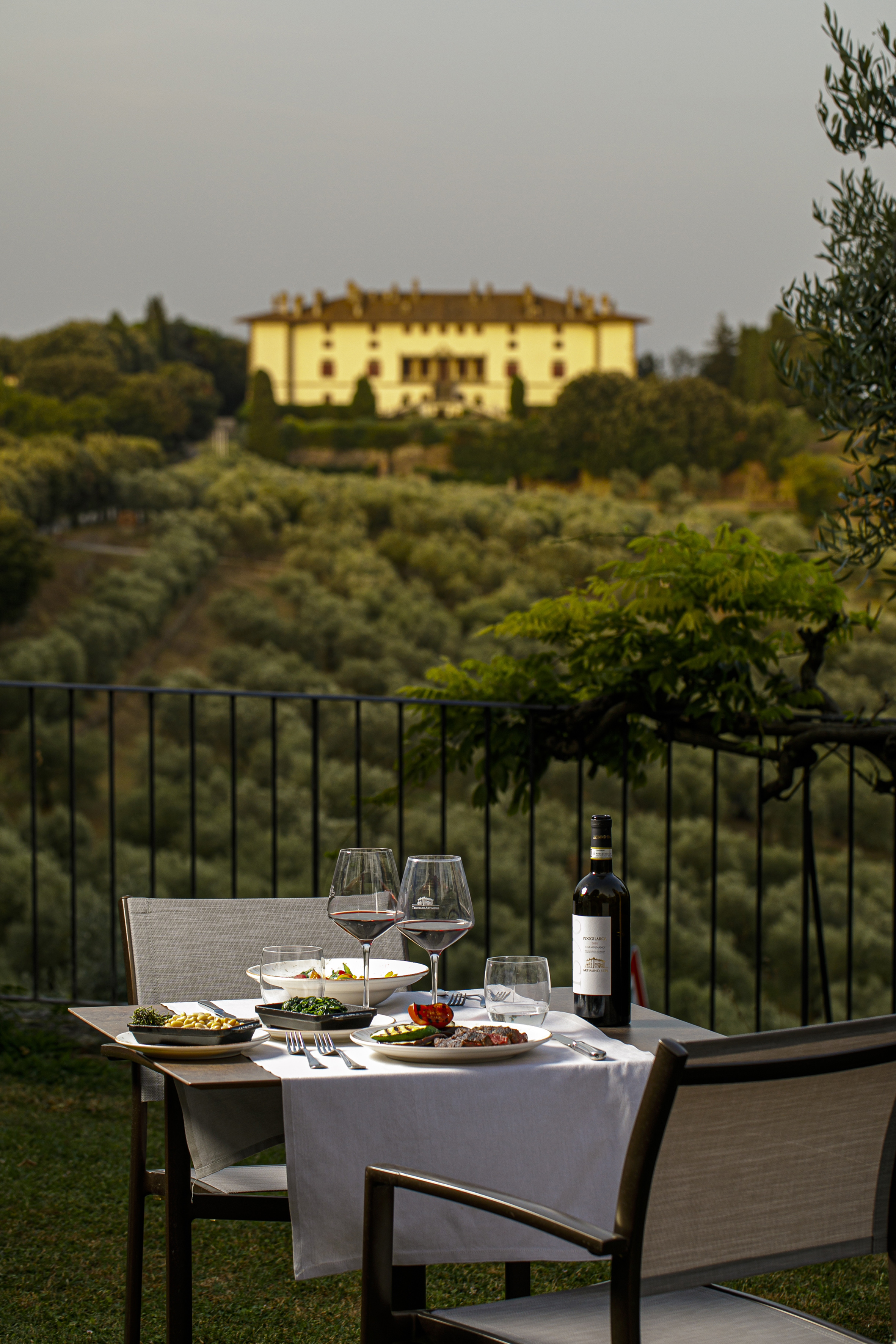 Outdoor table with red wine, food, overlooking Tuscan villa and olive groves.