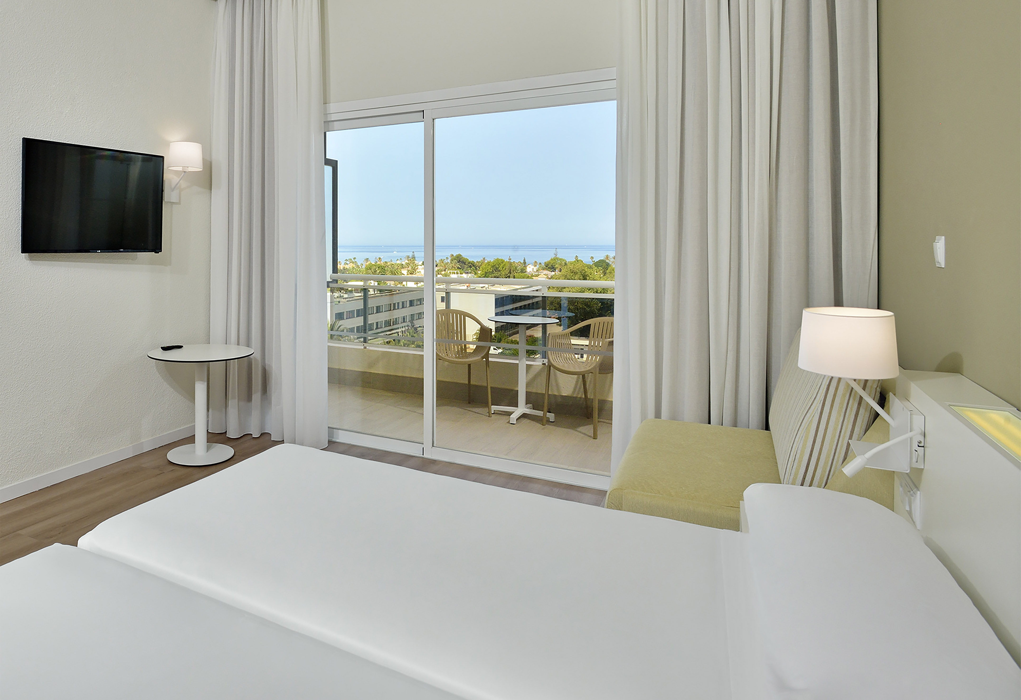 Bright hotel room with balcony, ocean view, twin beds, and TV.