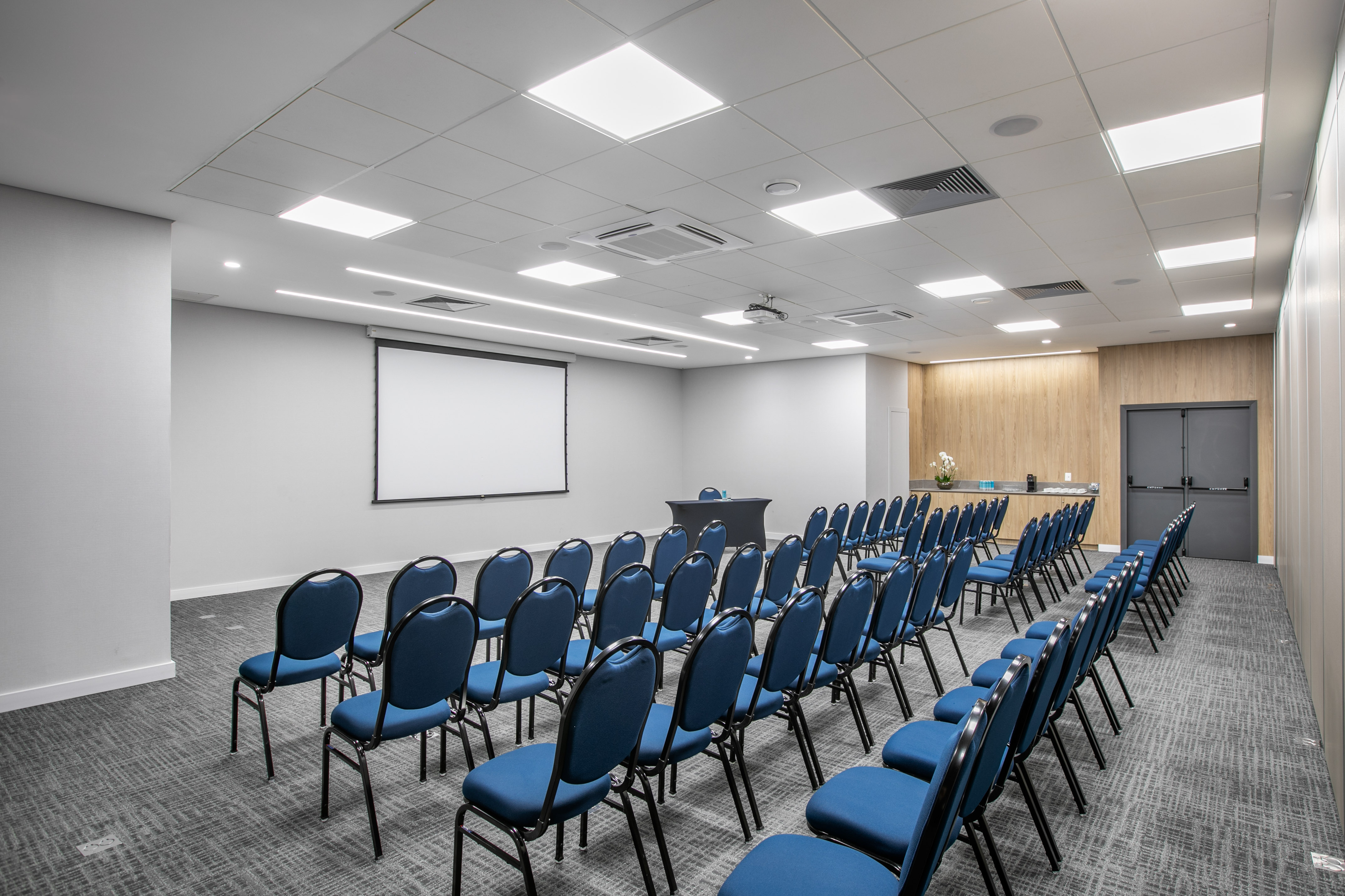 Conference room with rows of blue chairs, projection screen, and presenter's table.