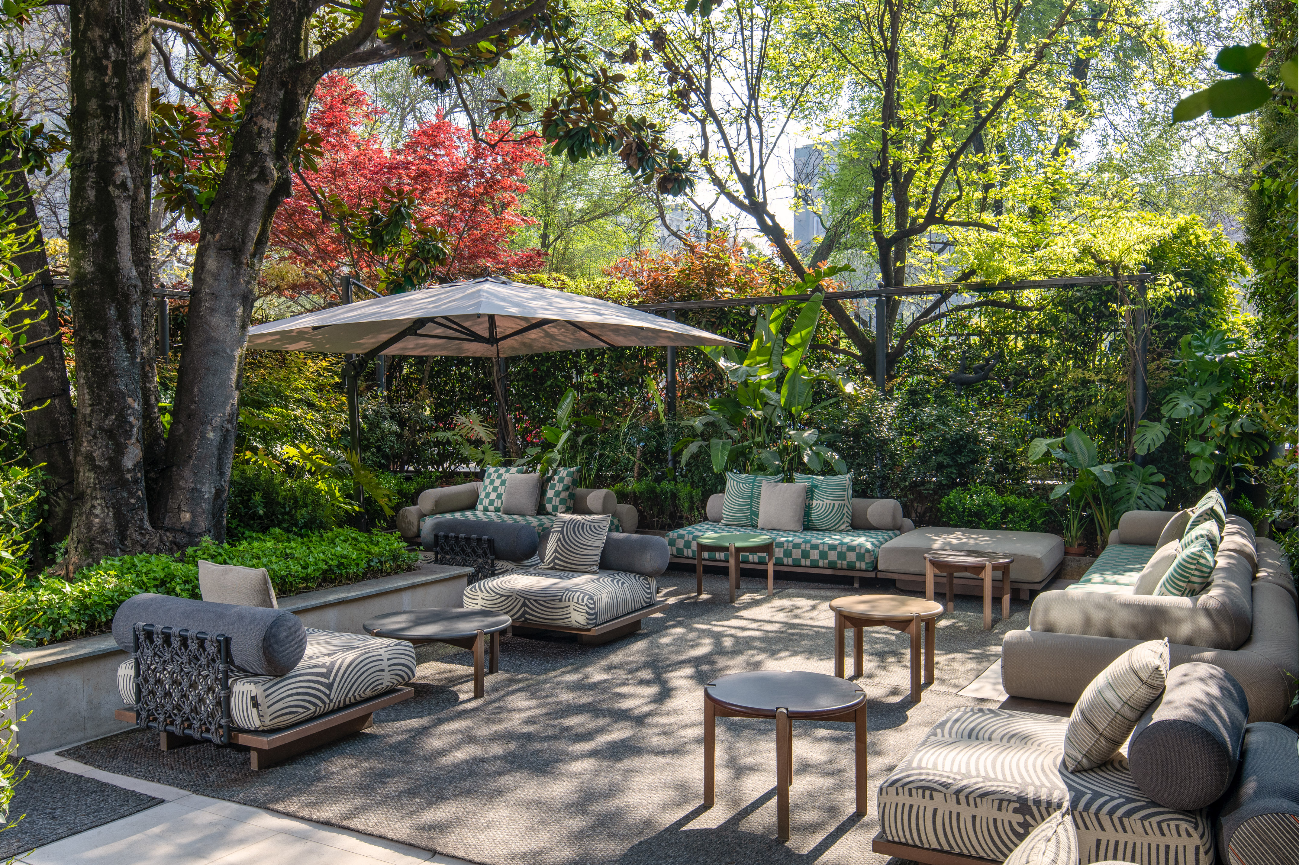 Lush garden patio with modern lounge furniture, tables, and a large sun umbrella.