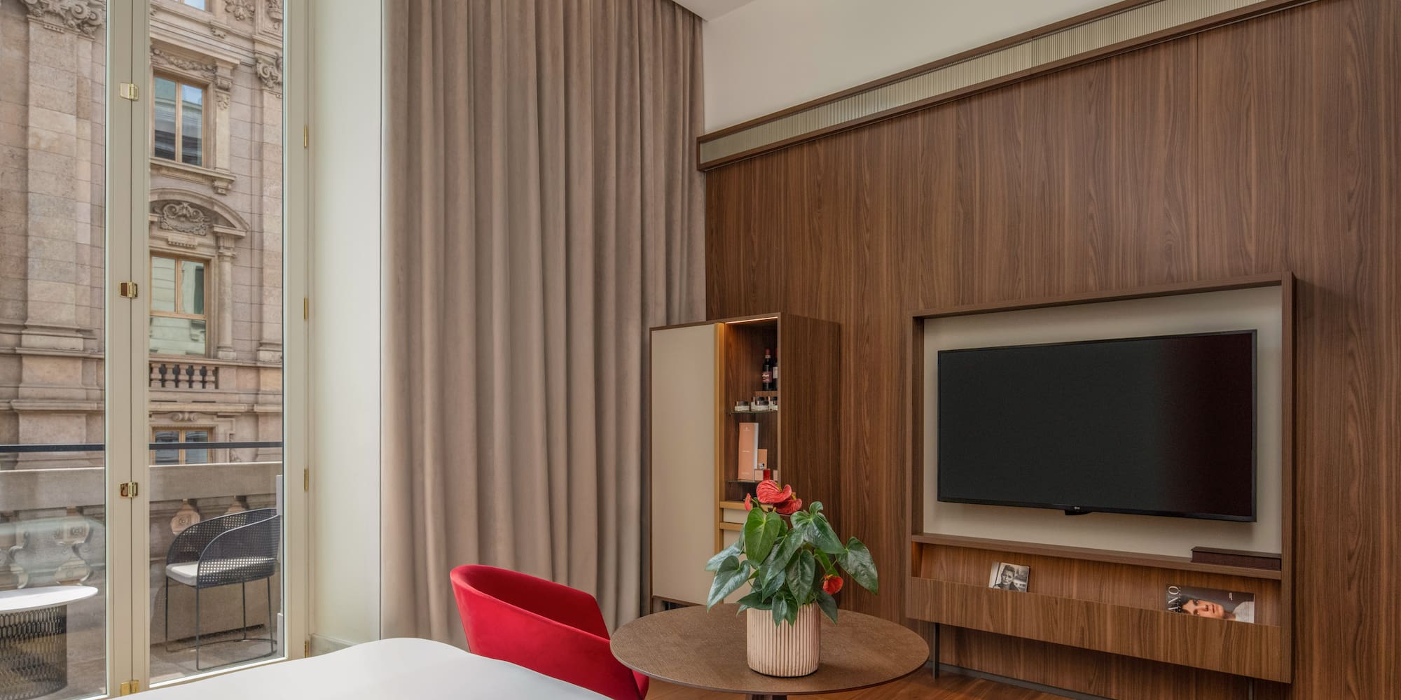Modern hotel room: wood wall, TV, red chairs, balcony with urban view.