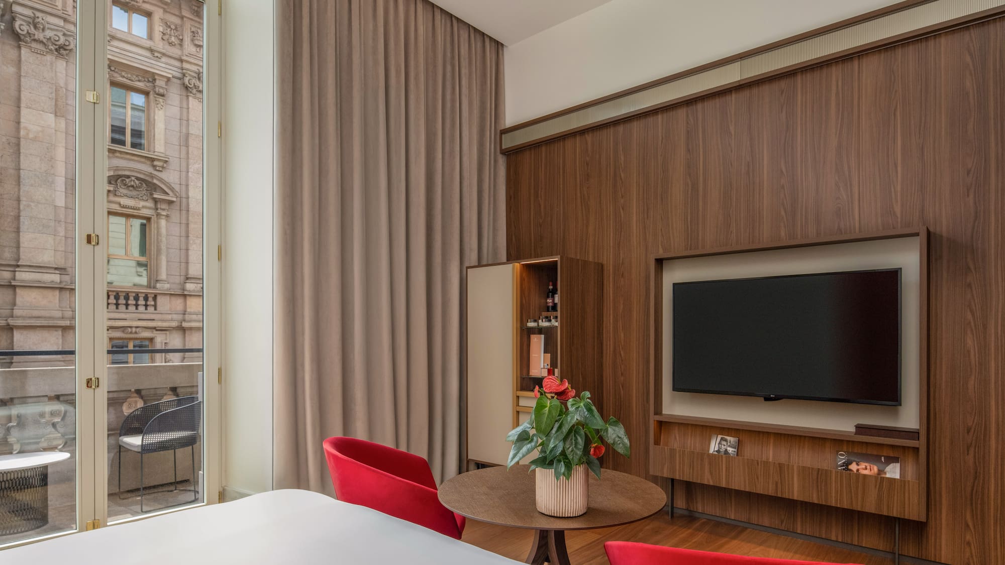 Modern hotel room: wood wall, TV, red chairs, balcony with urban view.