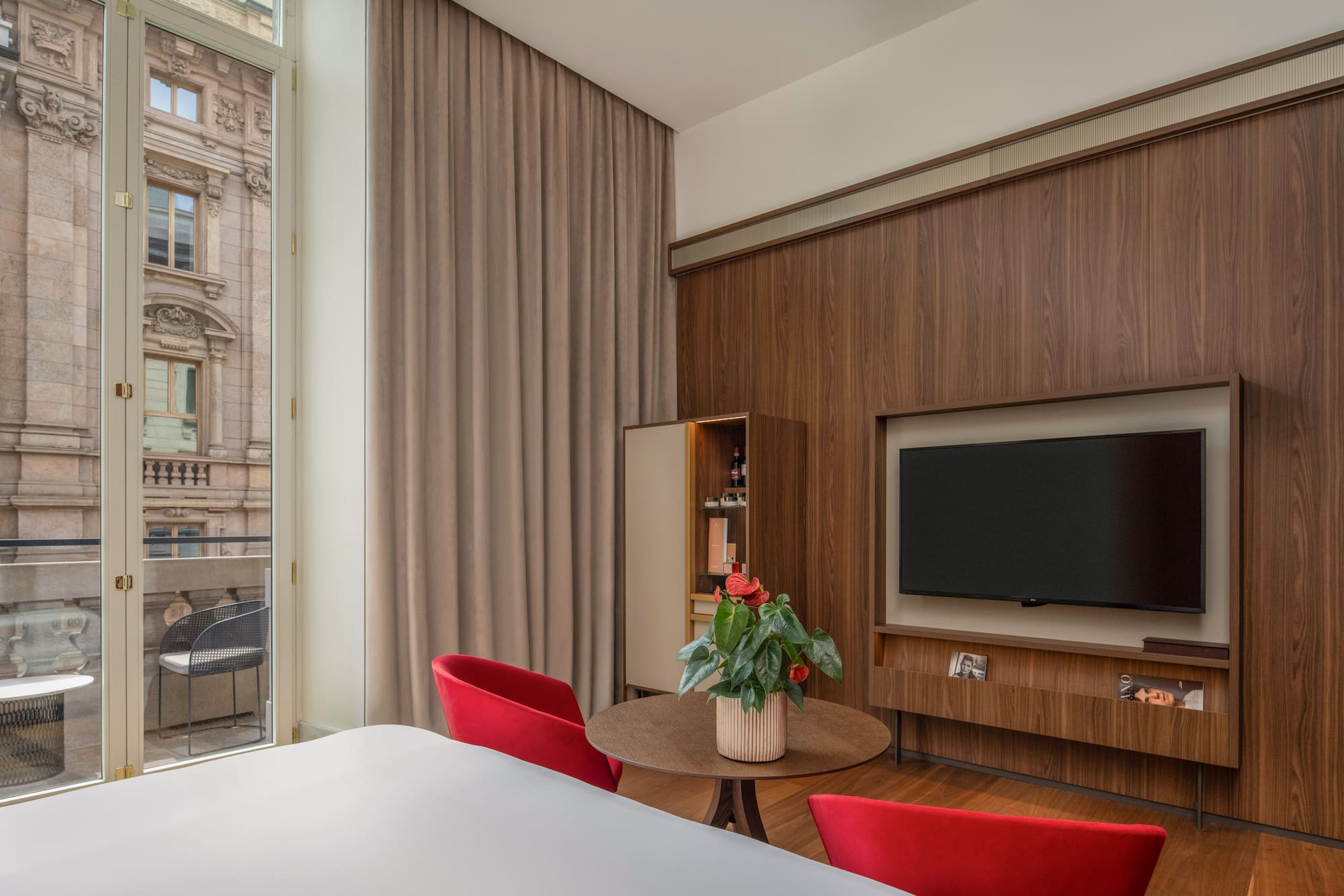 Modern hotel room: wood wall, TV, red chairs, balcony with urban view.