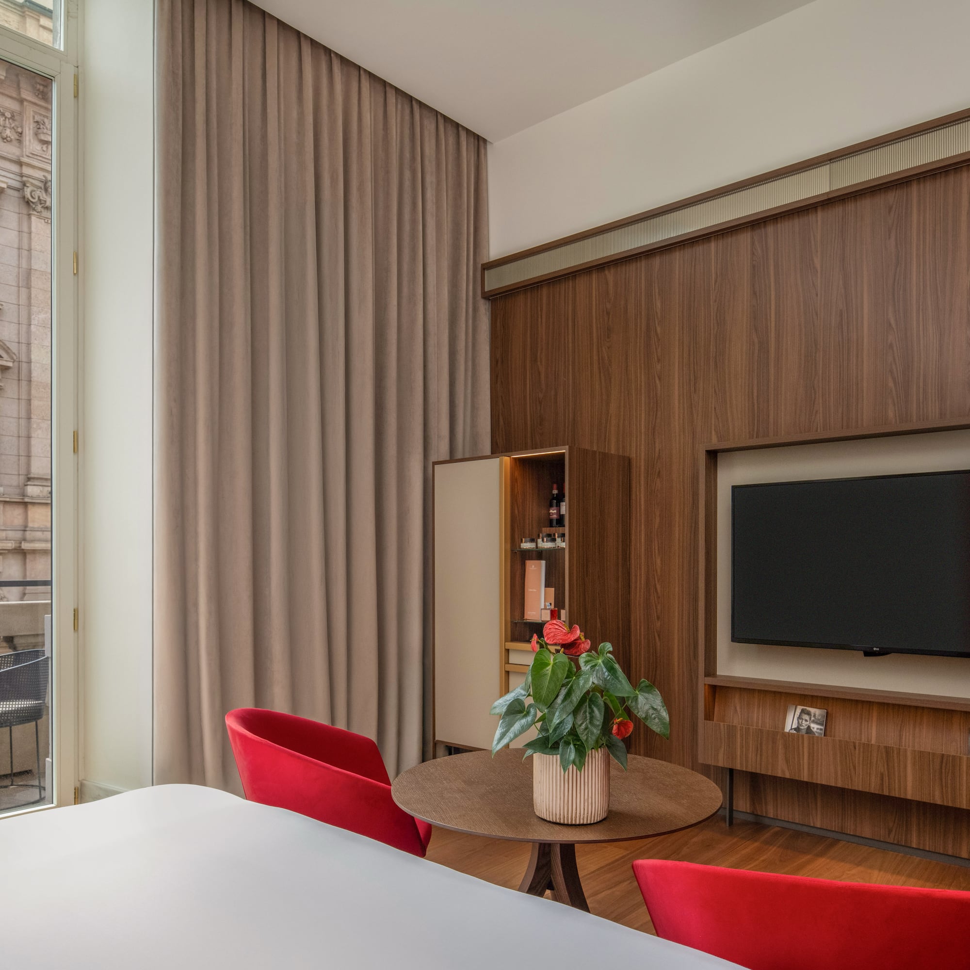 Modern hotel room: wood wall, TV, red chairs, balcony with urban view.