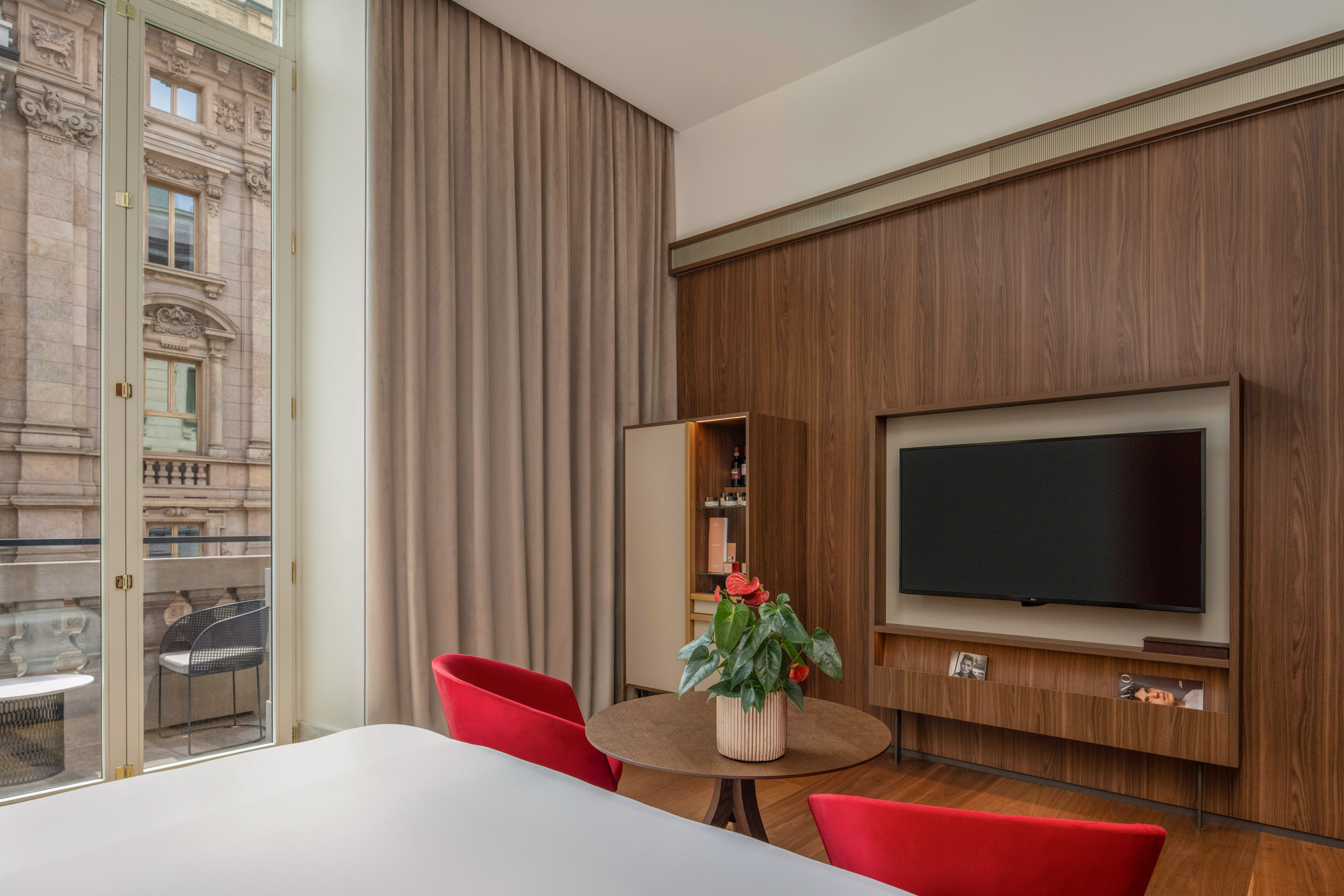 Modern hotel room: wood wall, TV, red chairs, balcony with urban view.