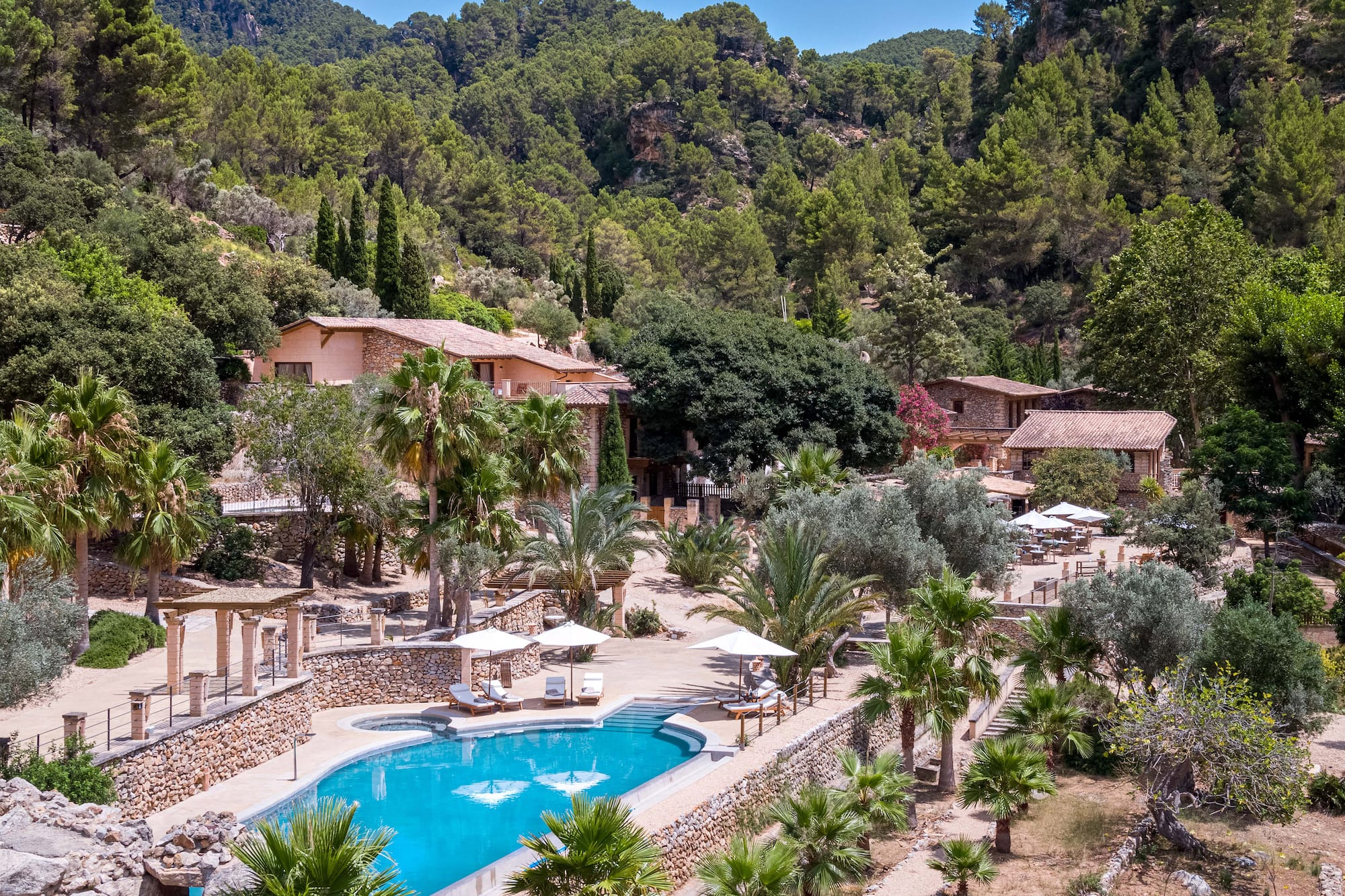 Luxury mountain resort with bright blue pool, palm trees, and stone buildings.