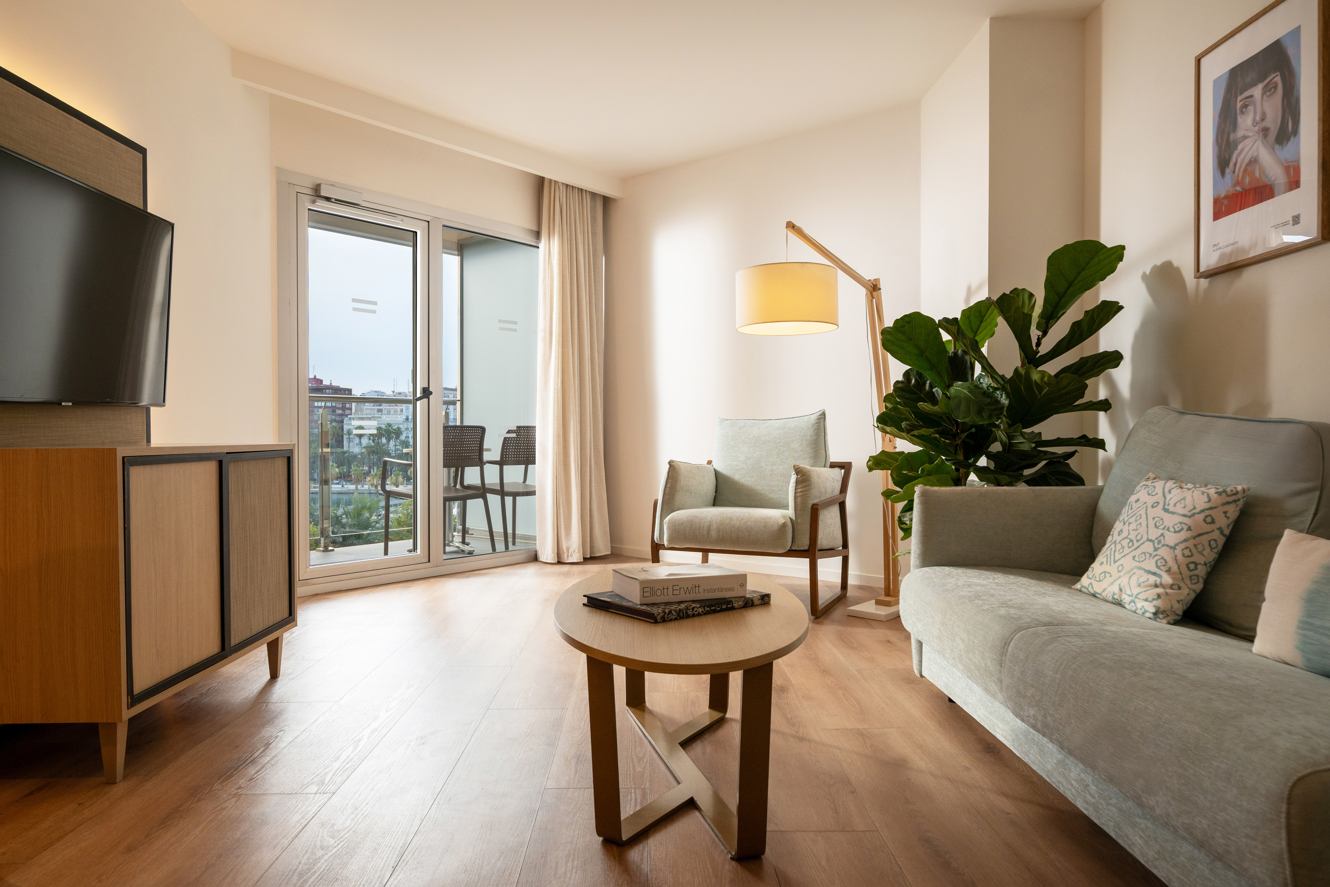 Modern hotel suite living room with sofa, coffee table, TV, plant, and balcony.