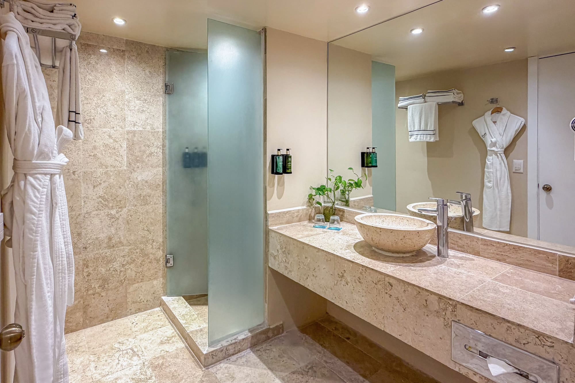 Modern hotel bathroom with walk-in shower, stone vanity, vessel sink, and white robes.