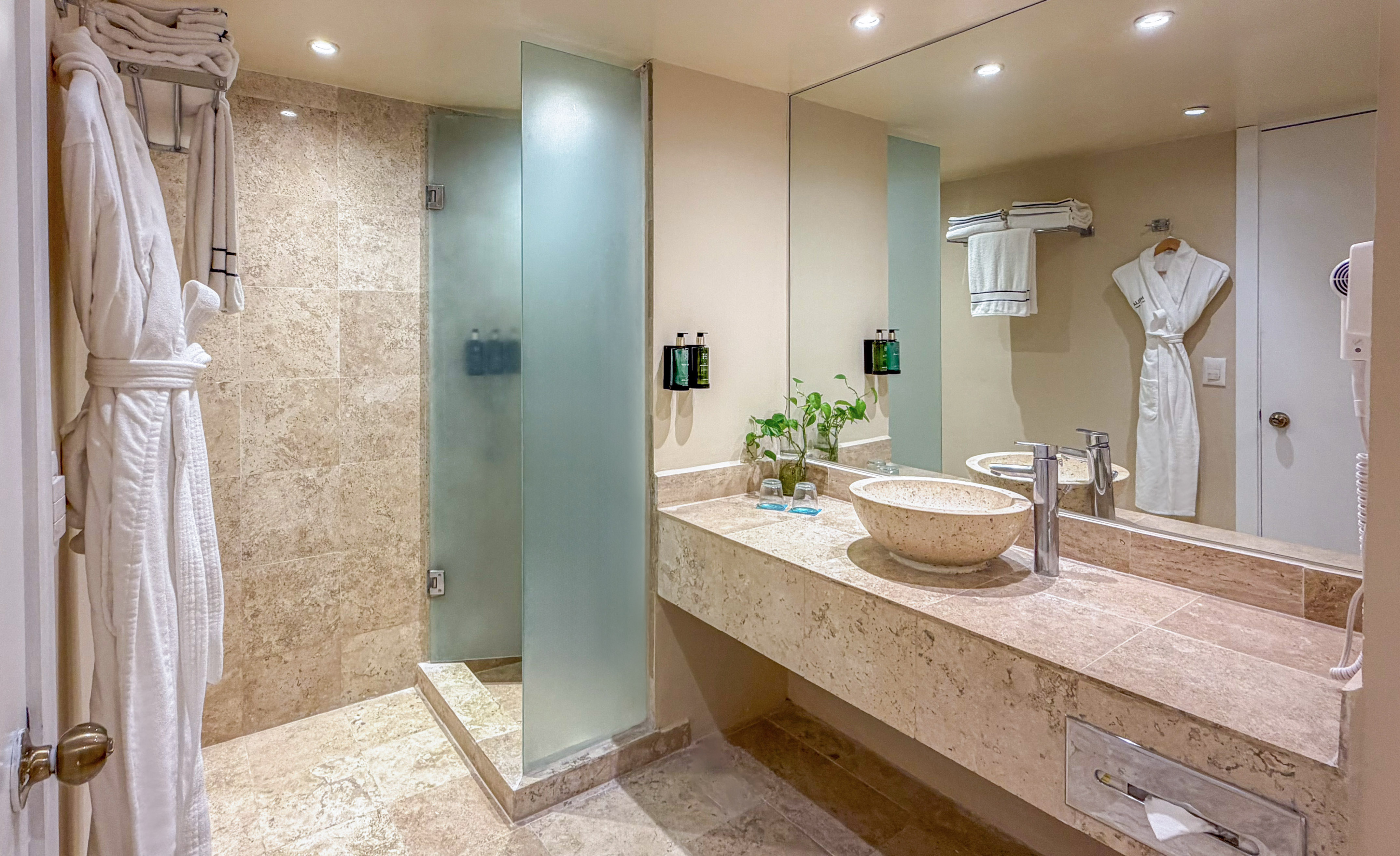 Modern hotel bathroom with walk-in shower, stone vanity, vessel sink, and white robes.