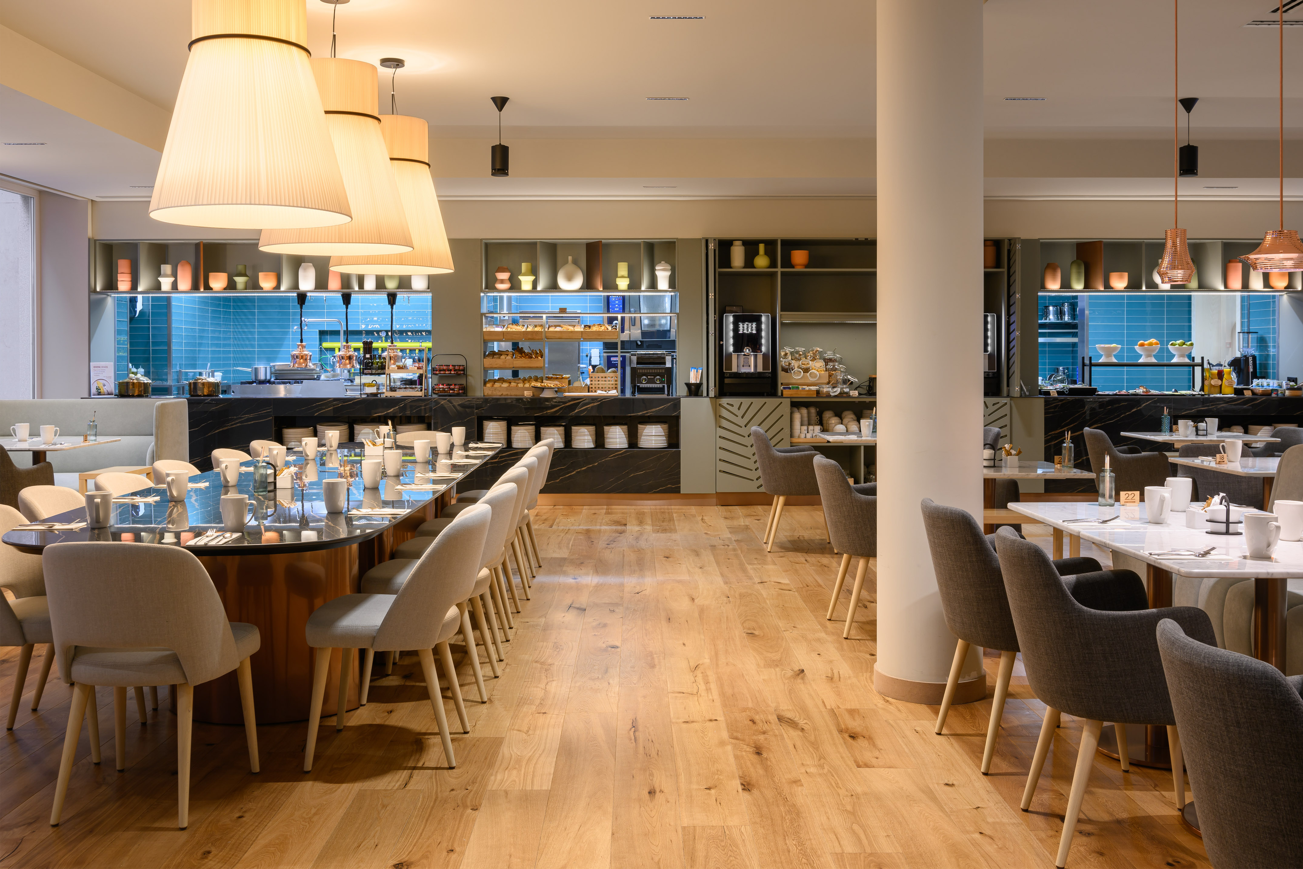 Modern hotel dining room with breakfast buffet, communal tables, and warm lighting.