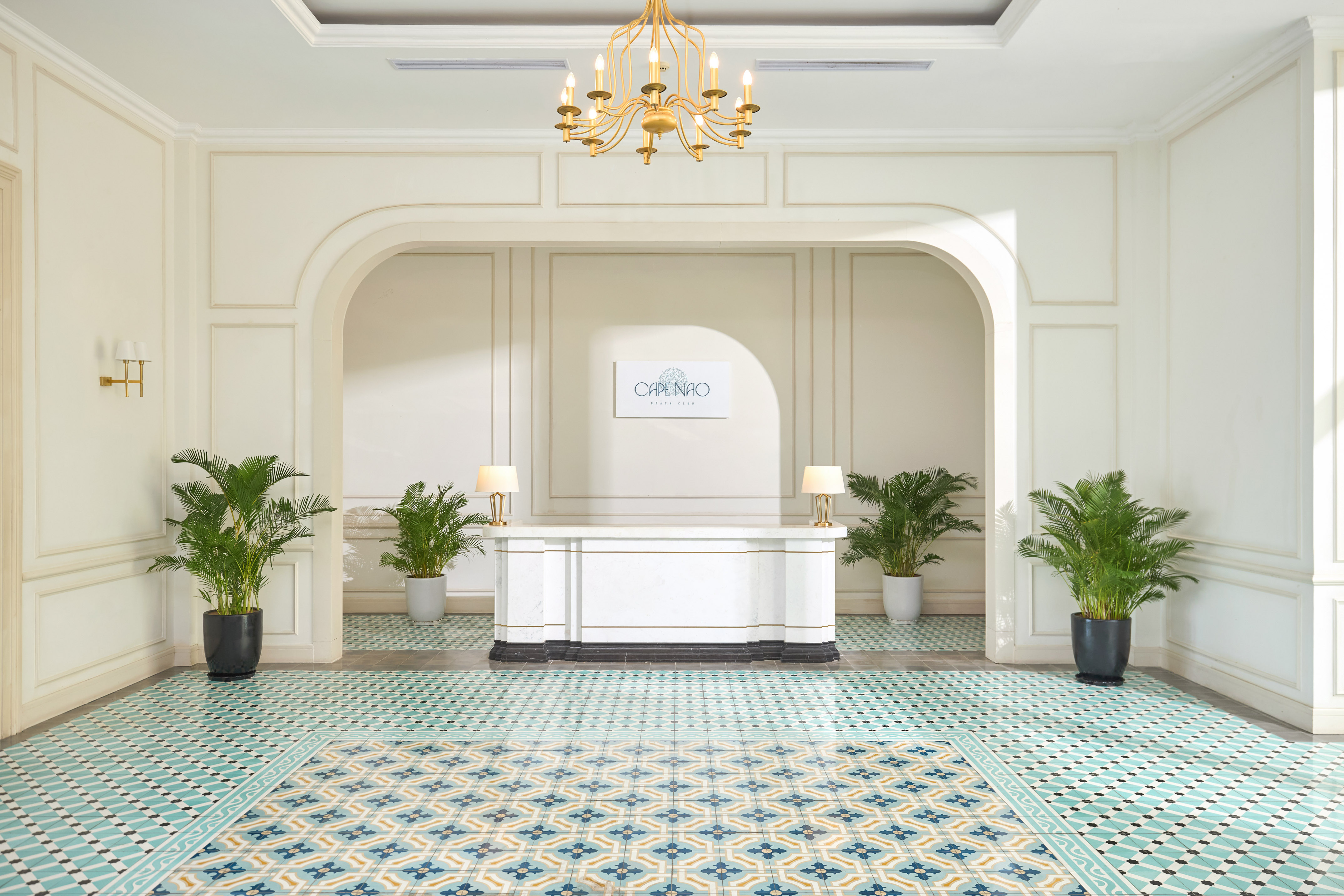 Elegant hotel reception with marble desk, patterned floor, and palm plants.