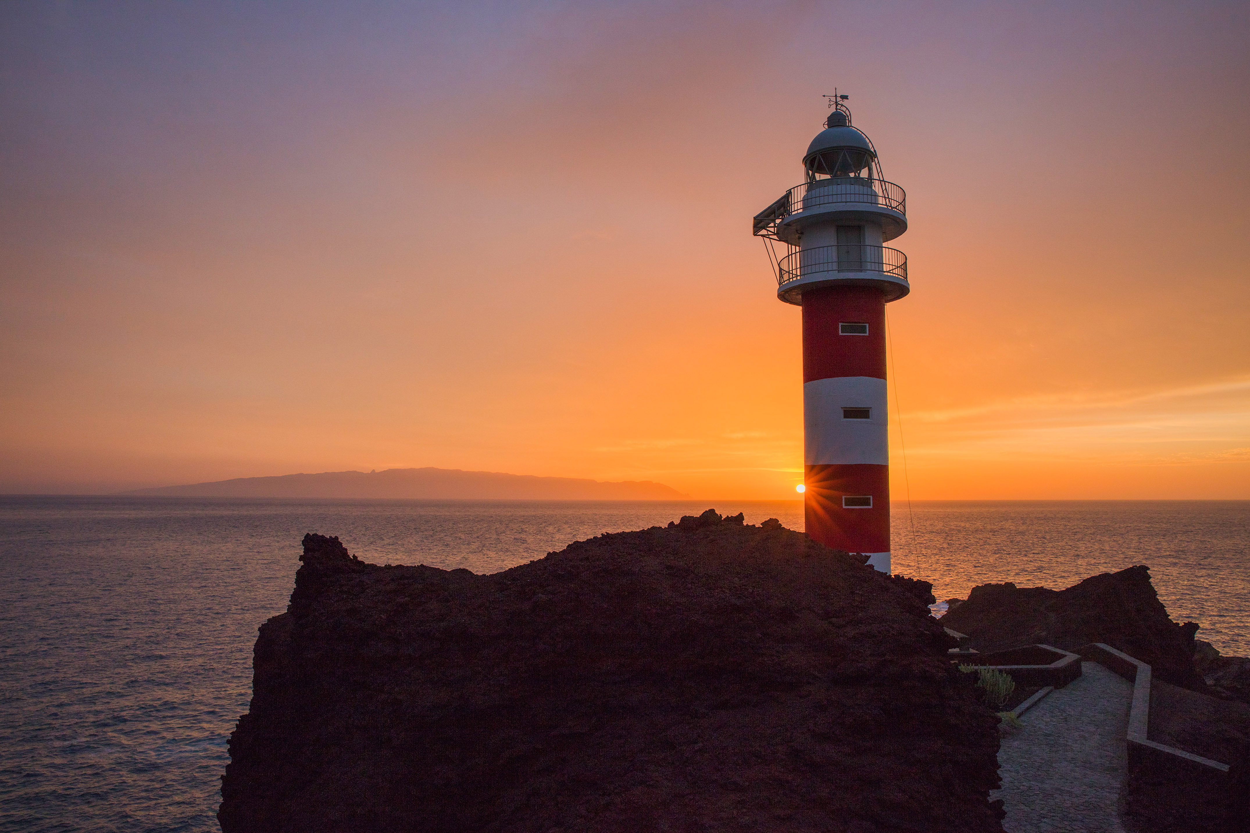 Faro rojo y blanco en acantilado rocoso al atardecer sobre el mar.