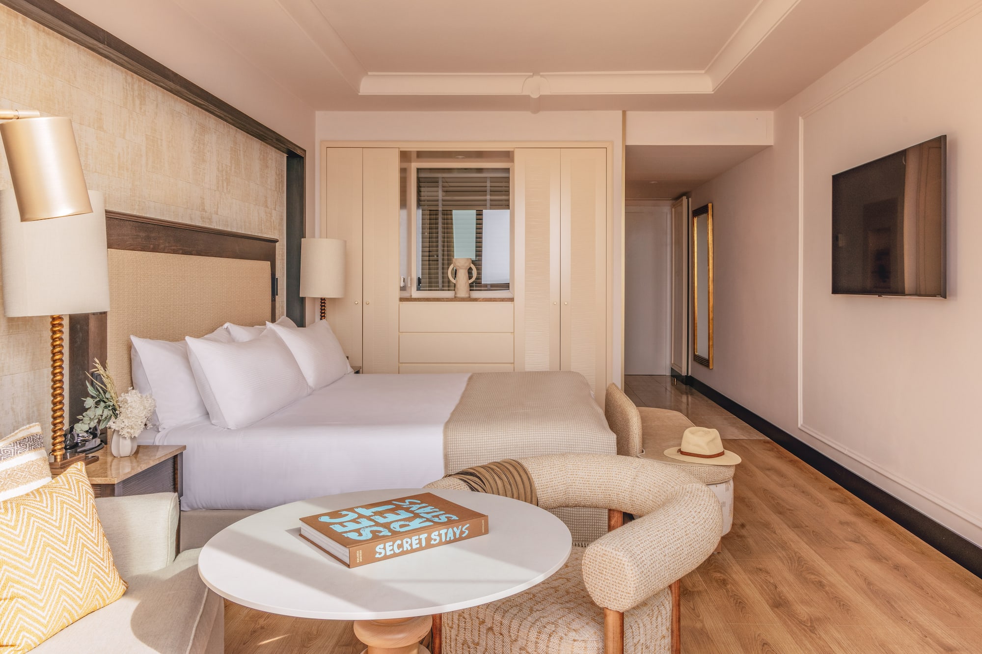 Modern hotel bedroom with king bed, neutral decor, seating area, and TV.