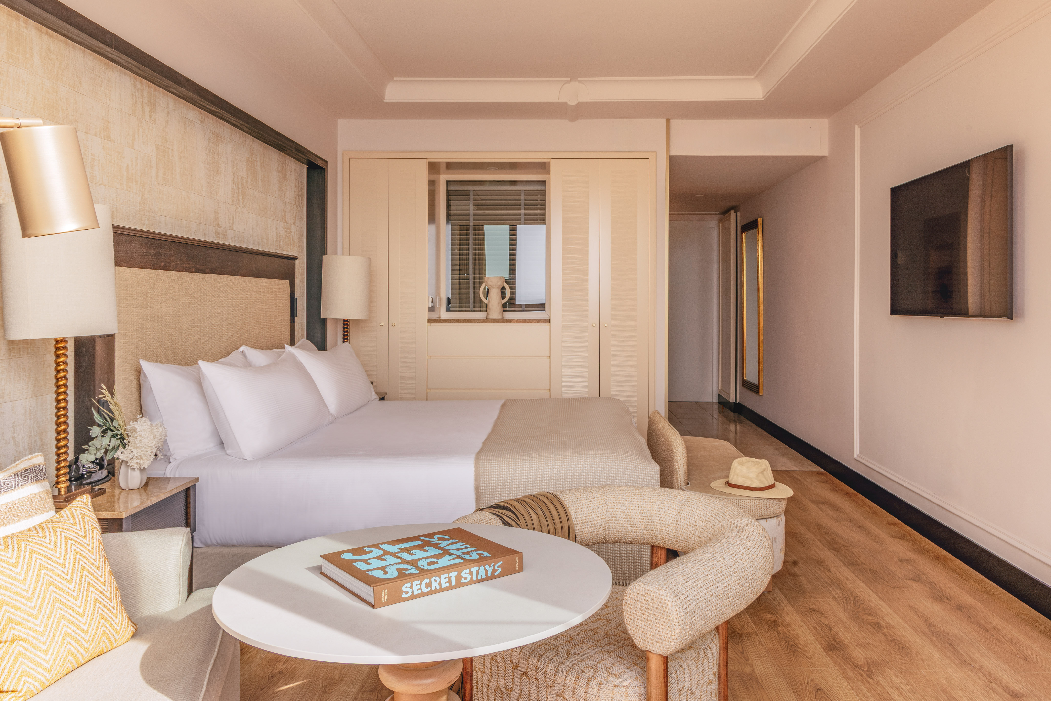 Modern hotel bedroom with king bed, neutral decor, seating area, and TV.