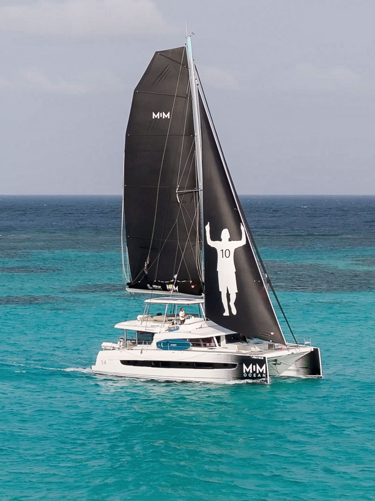 Catamarán MIM Ocean con vela negra Messi 10 en aguas turquesas.