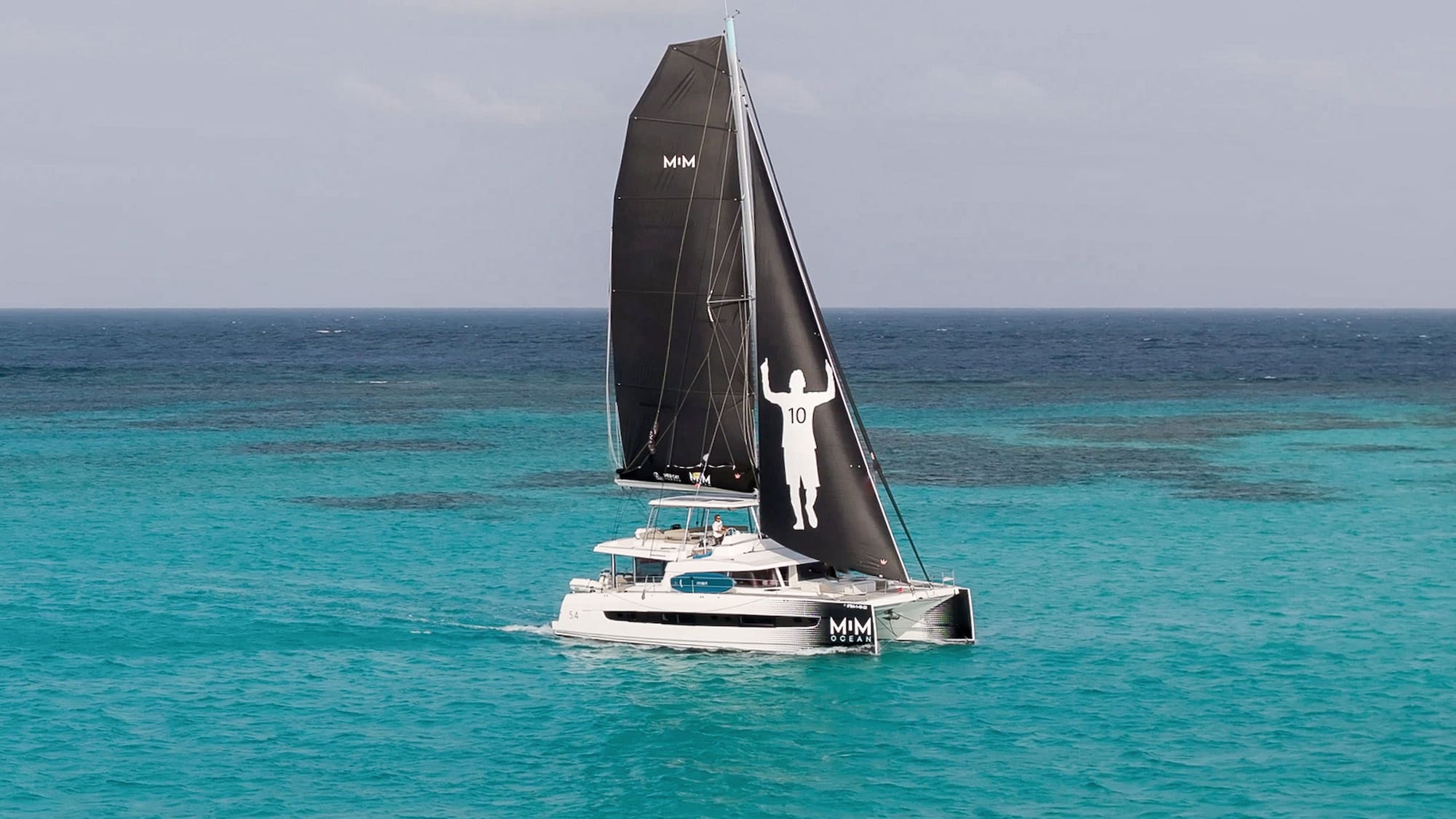 Catamaran with black Messi 10 sails on clear turquoise water.