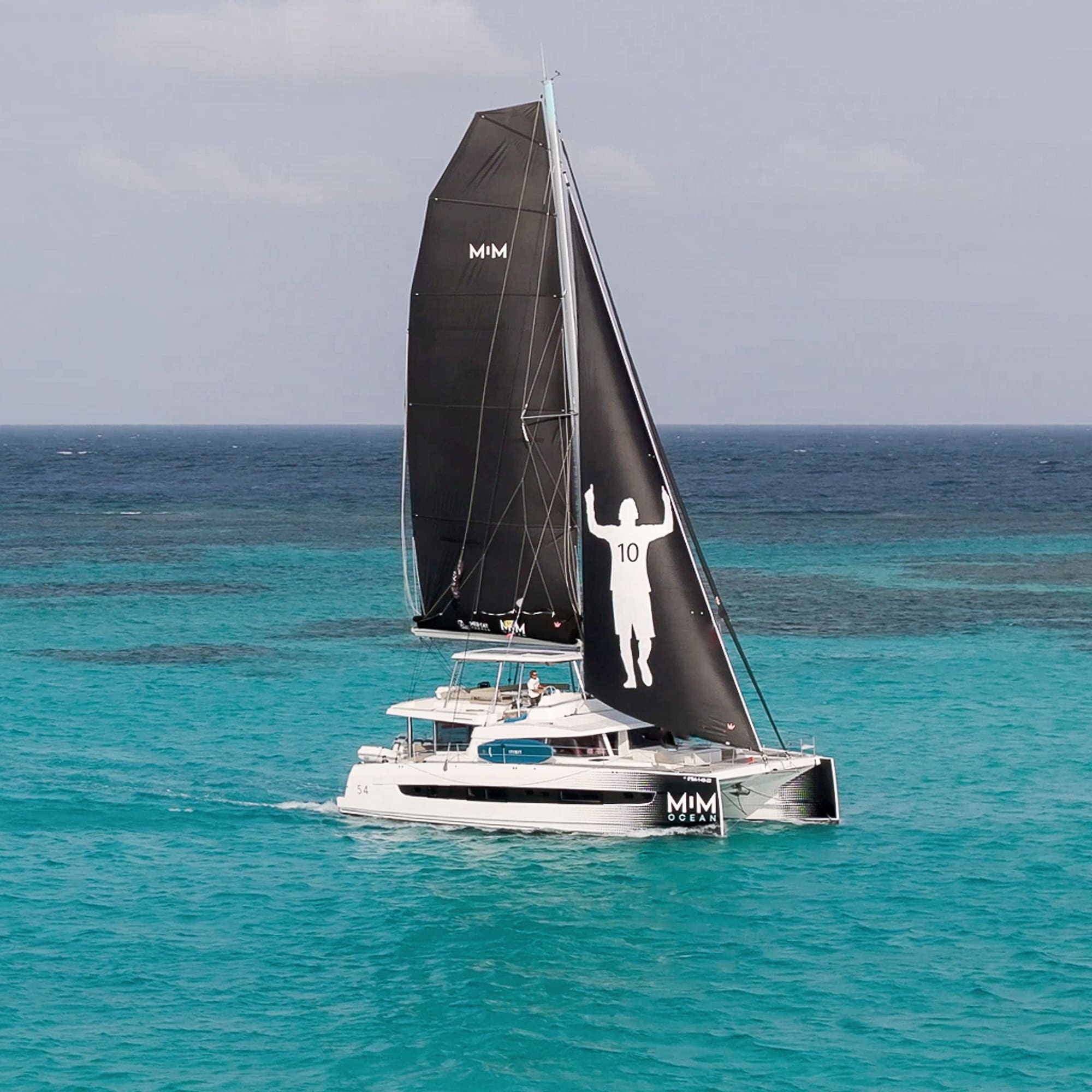 Catamarán MIM Ocean con vela negra Messi 10 en aguas turquesas.