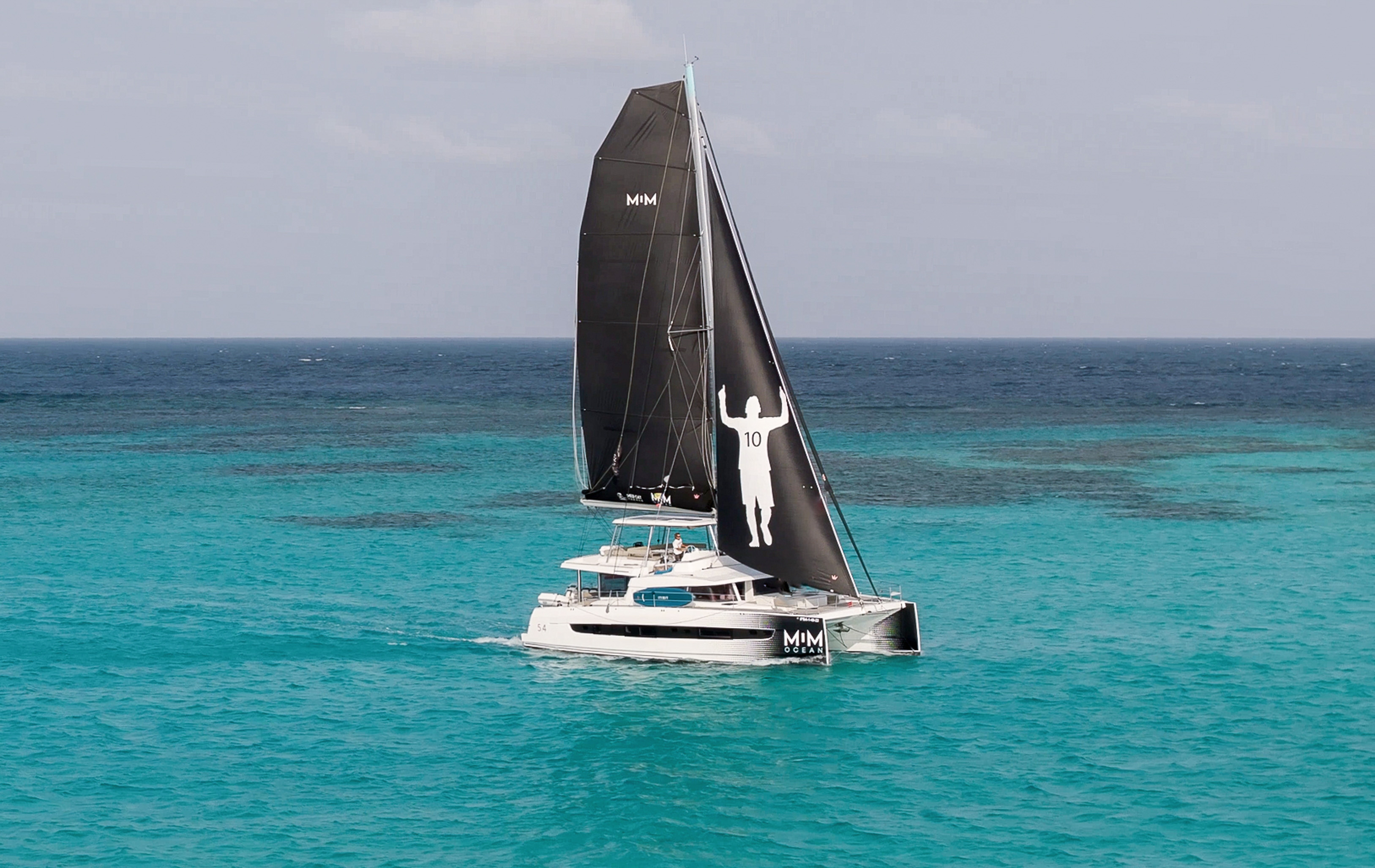 Catamaran with black Messi 10 sails on clear turquoise water.