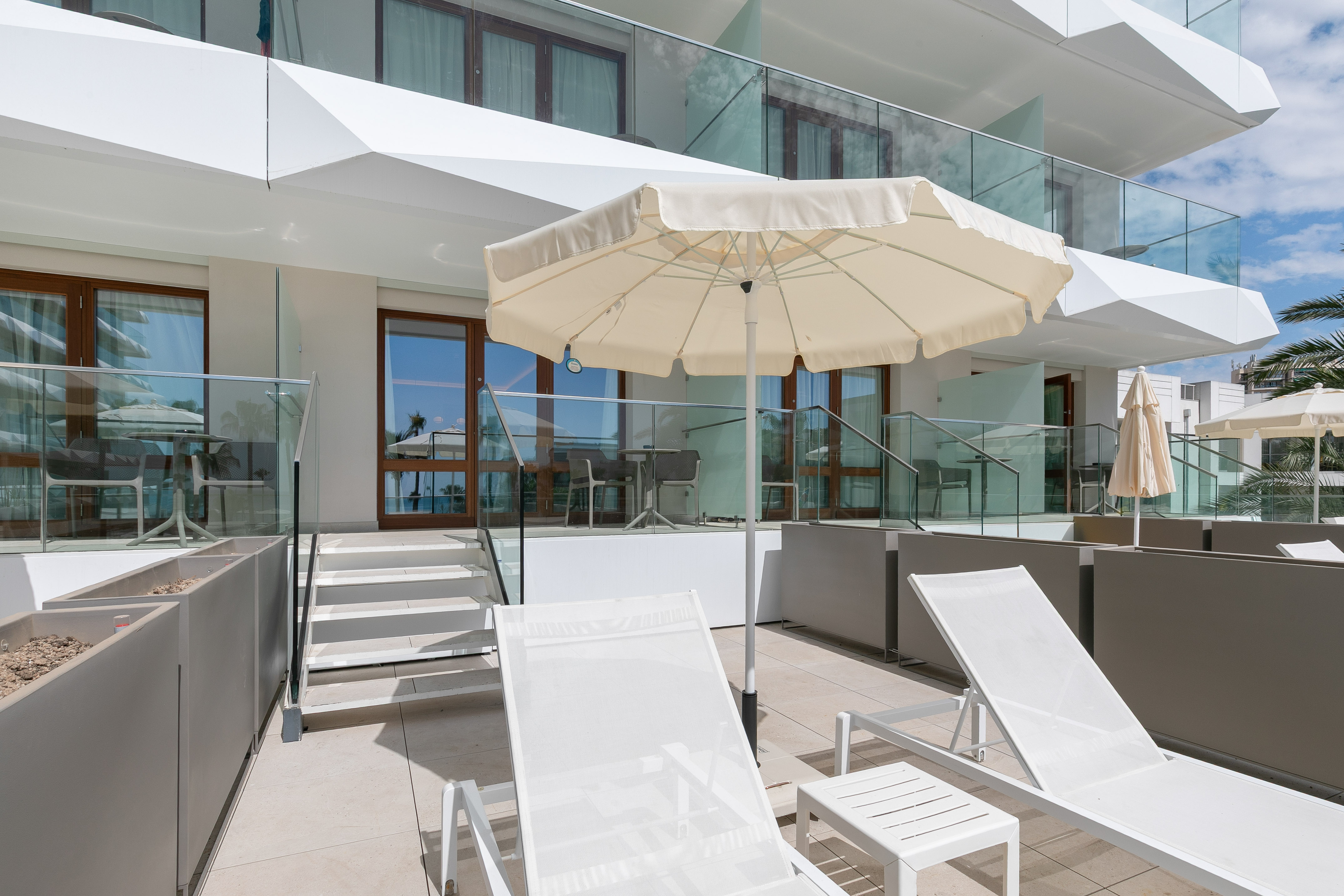 Modern resort patio with white sun loungers and beige umbrella.