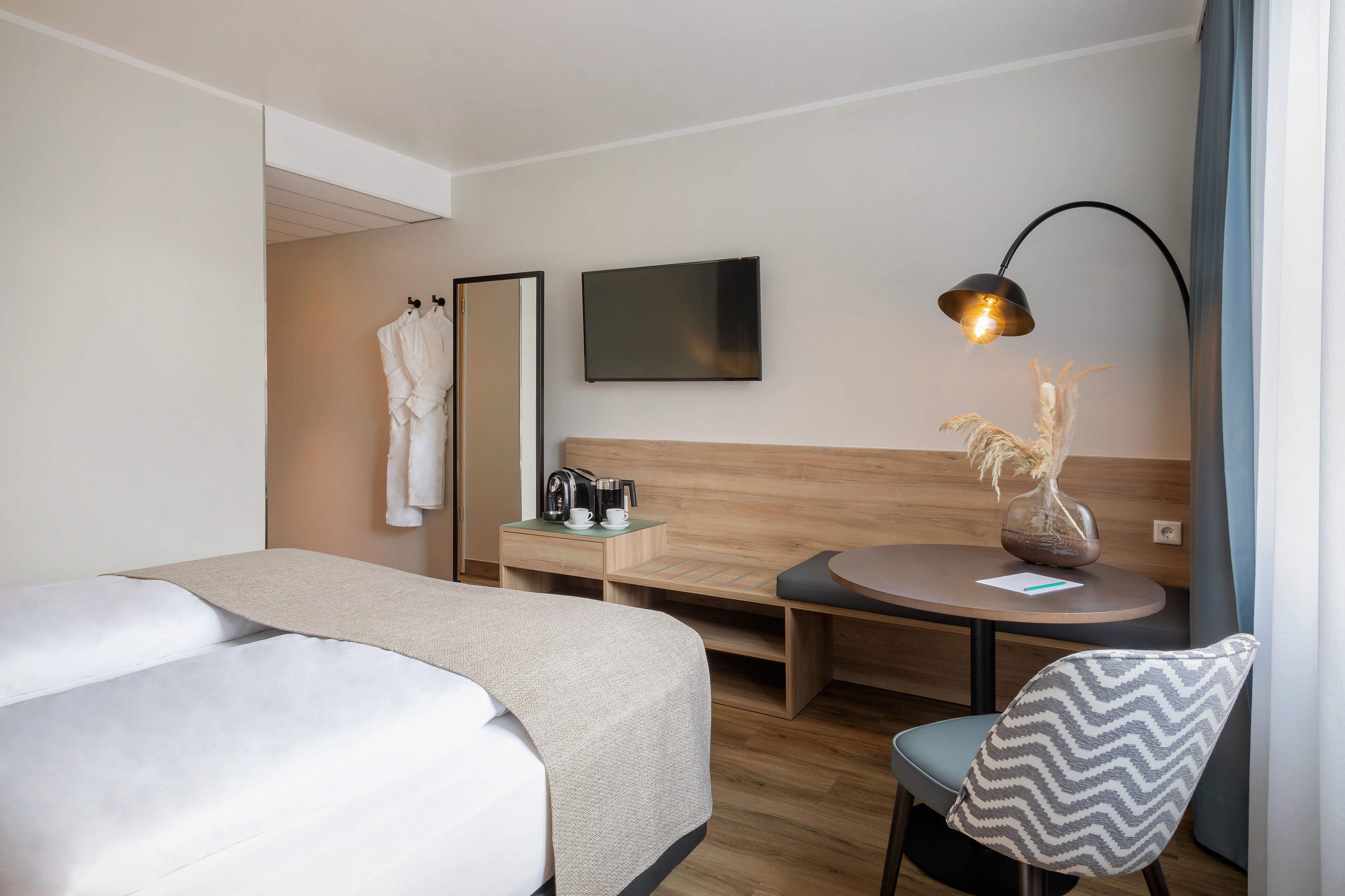 Modern hotel room with bed, TV, coffee station, round table, and chevron chair.
