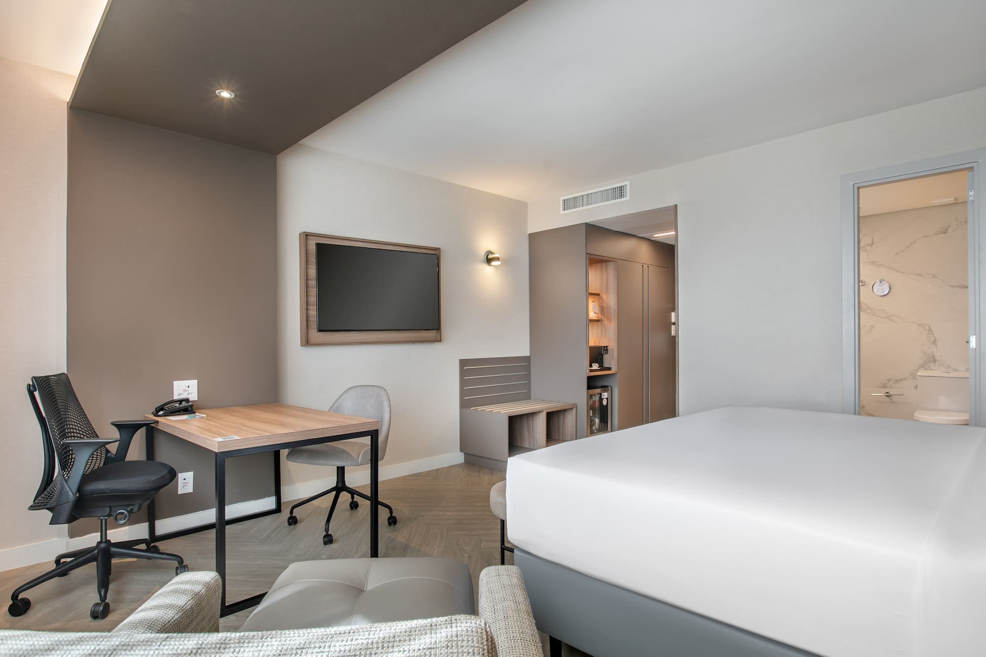 Contemporary hotel room with bed, work desk, wall TV, and mini-bar.