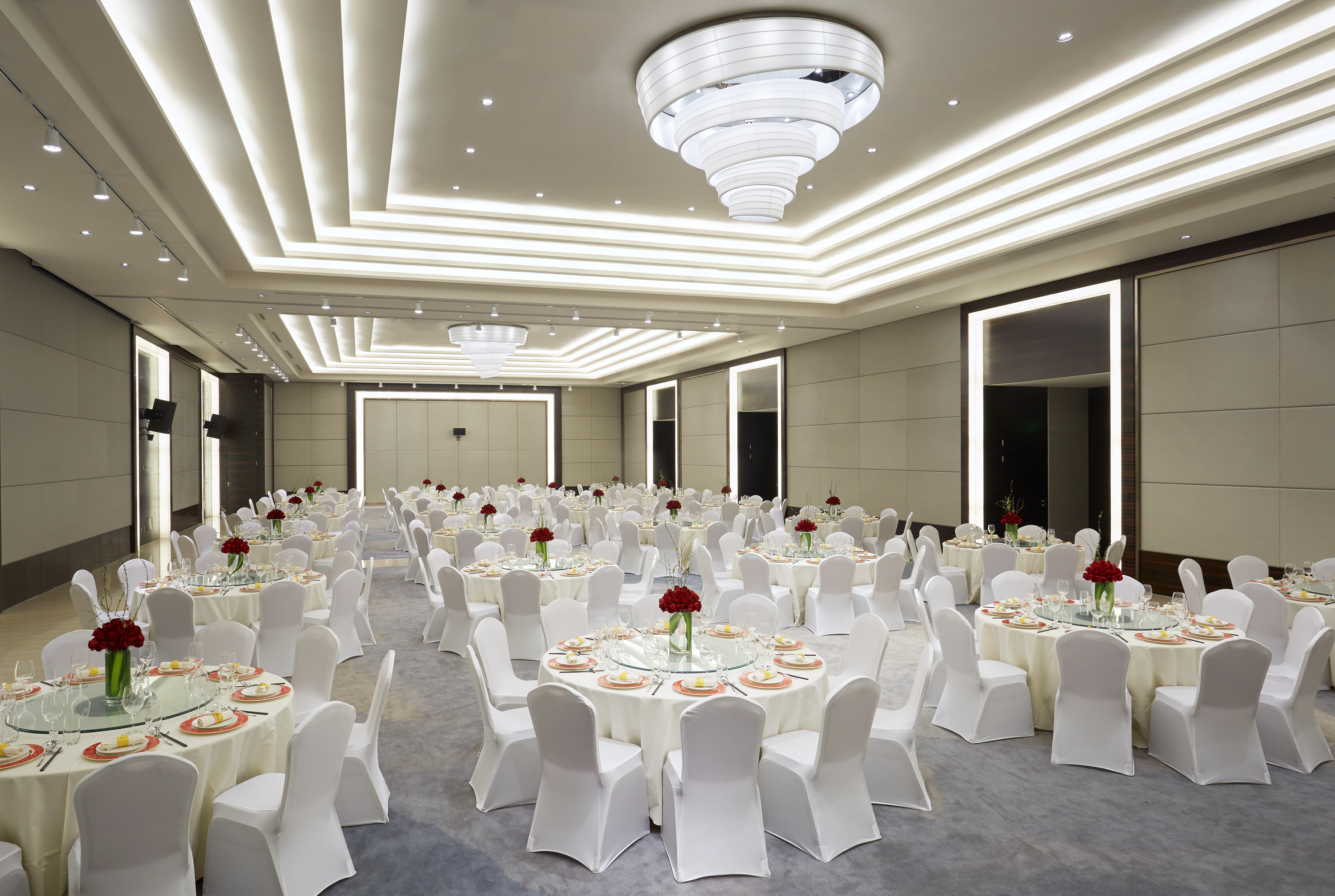 Elegant ballroom with round tables, white chairs, red centerpieces, modern chandeliers.