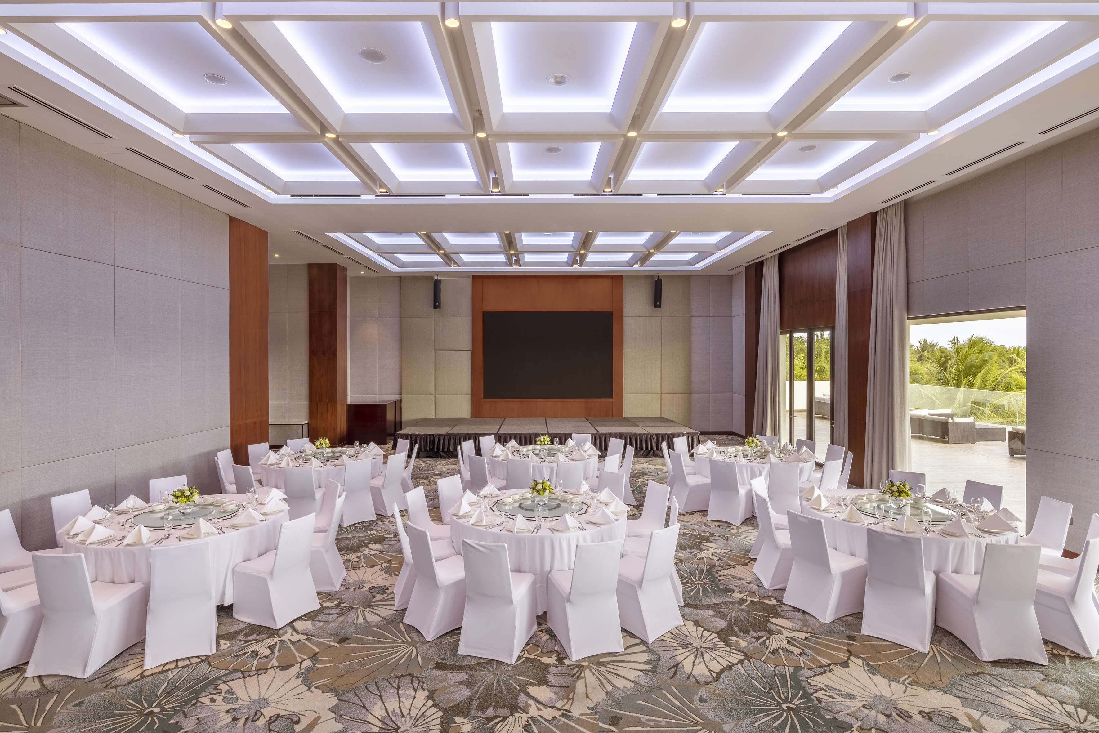 Modern banquet hall with dining tables, stage, screen, and tropical view.