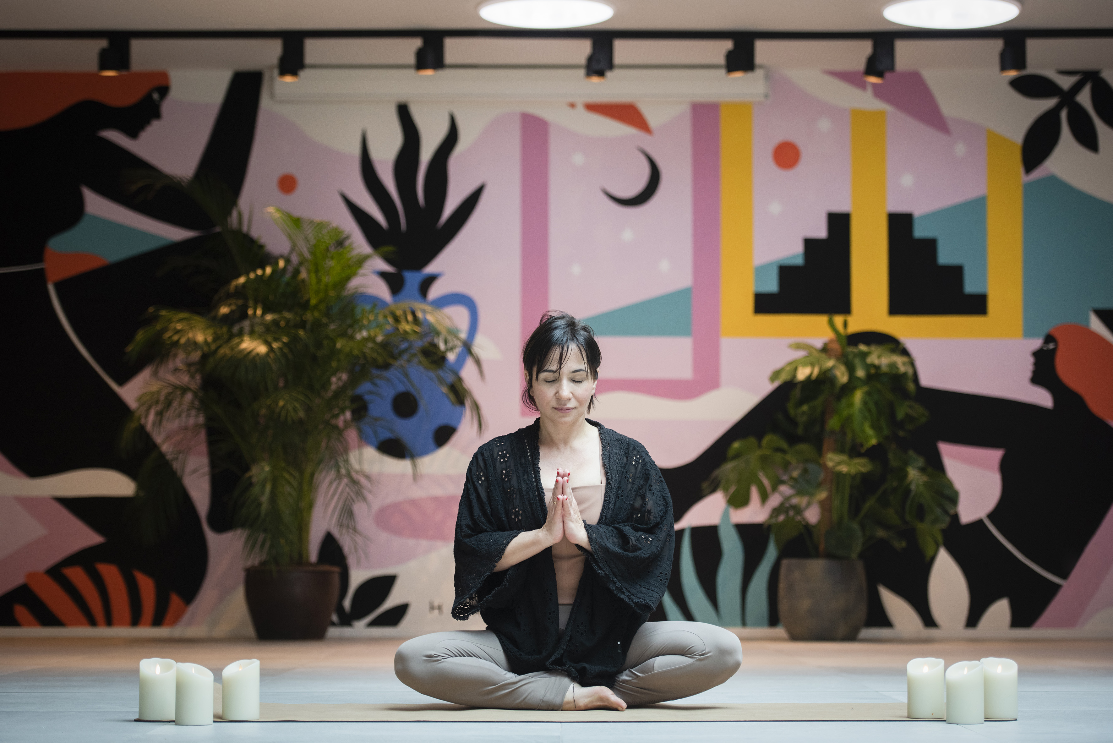 Woman meditating in lotus pose on yoga mat, colorful studio mural, candles.