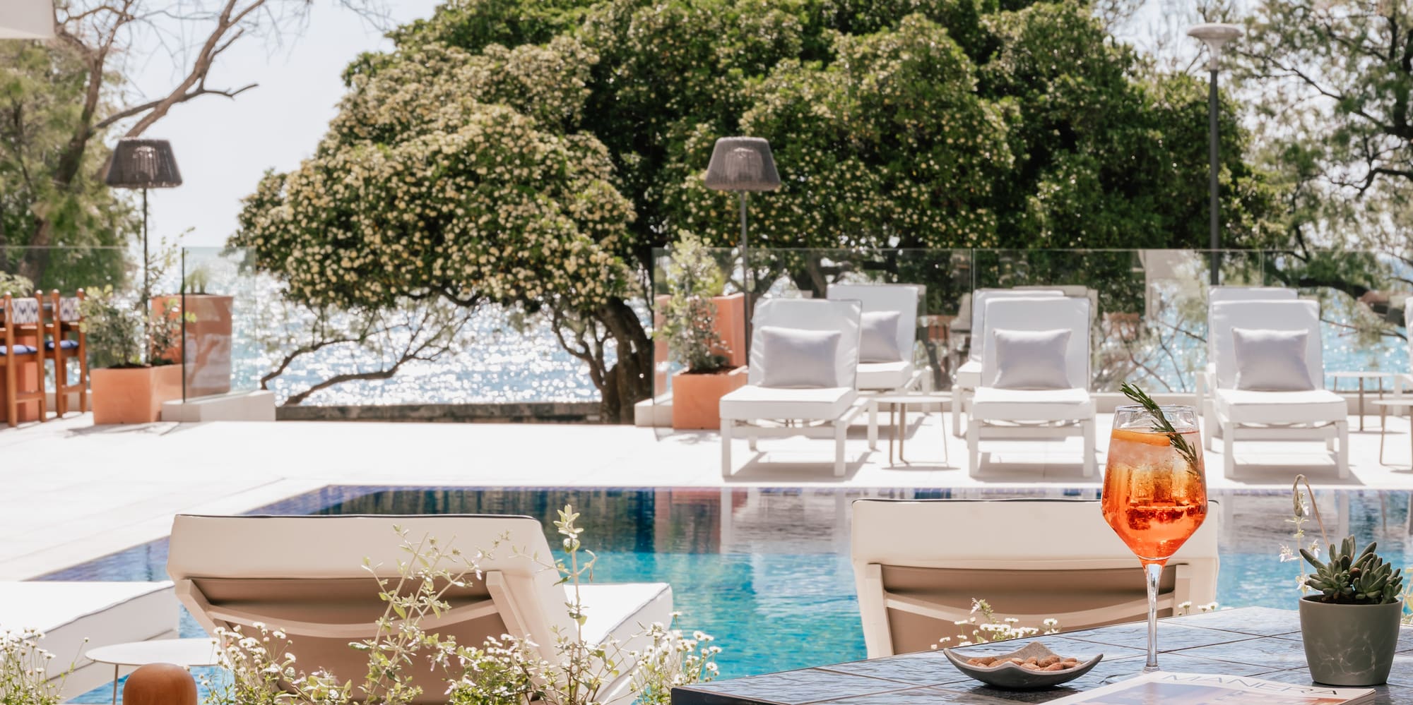 Orange cocktail on table by resort pool, sun loungers, and sparkling ocean.