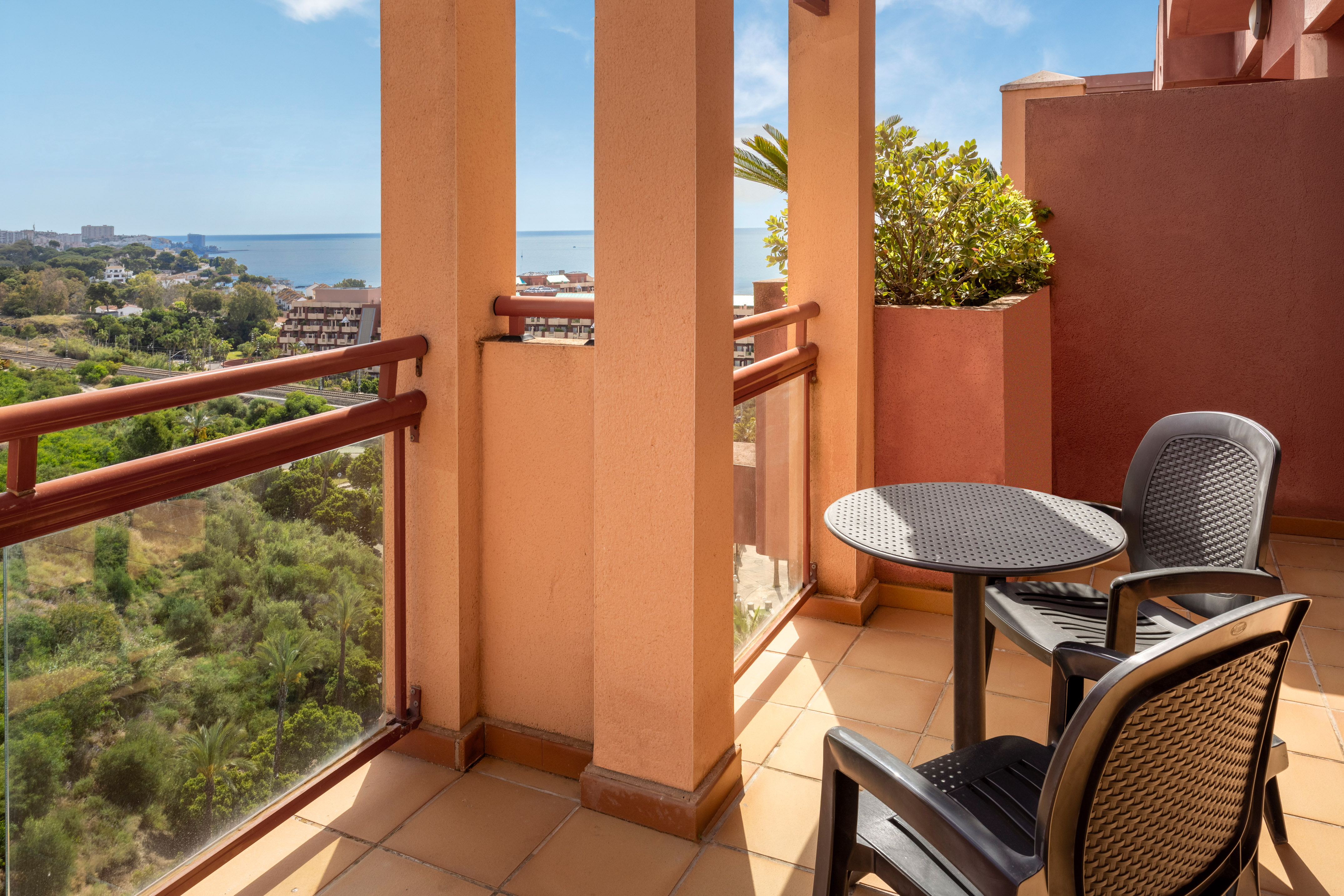 Terracotta hotel balcony with sea view, outdoor furniture, and lush greenery.