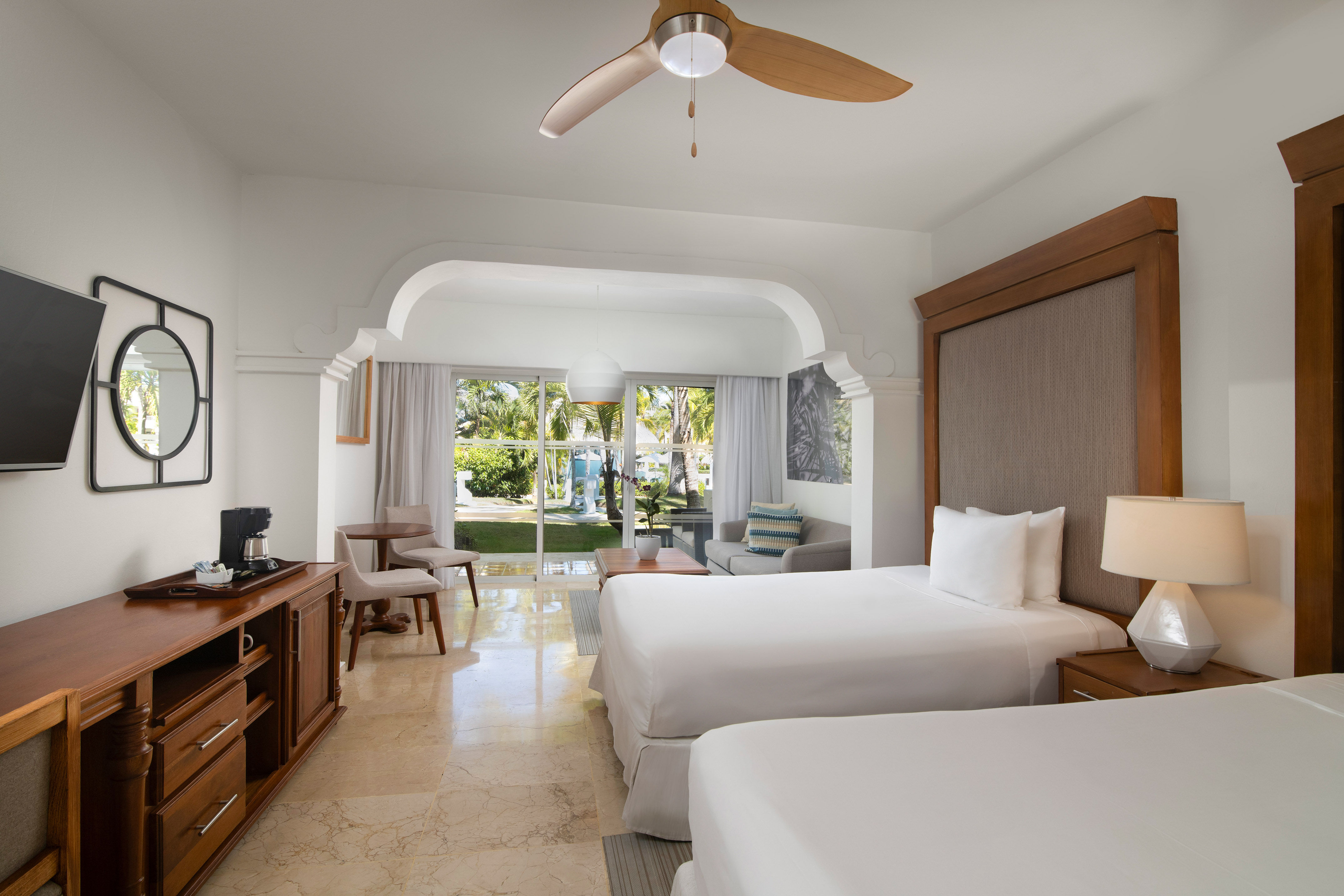 Bright hotel room with two beds, sofa, and tropical garden patio view.