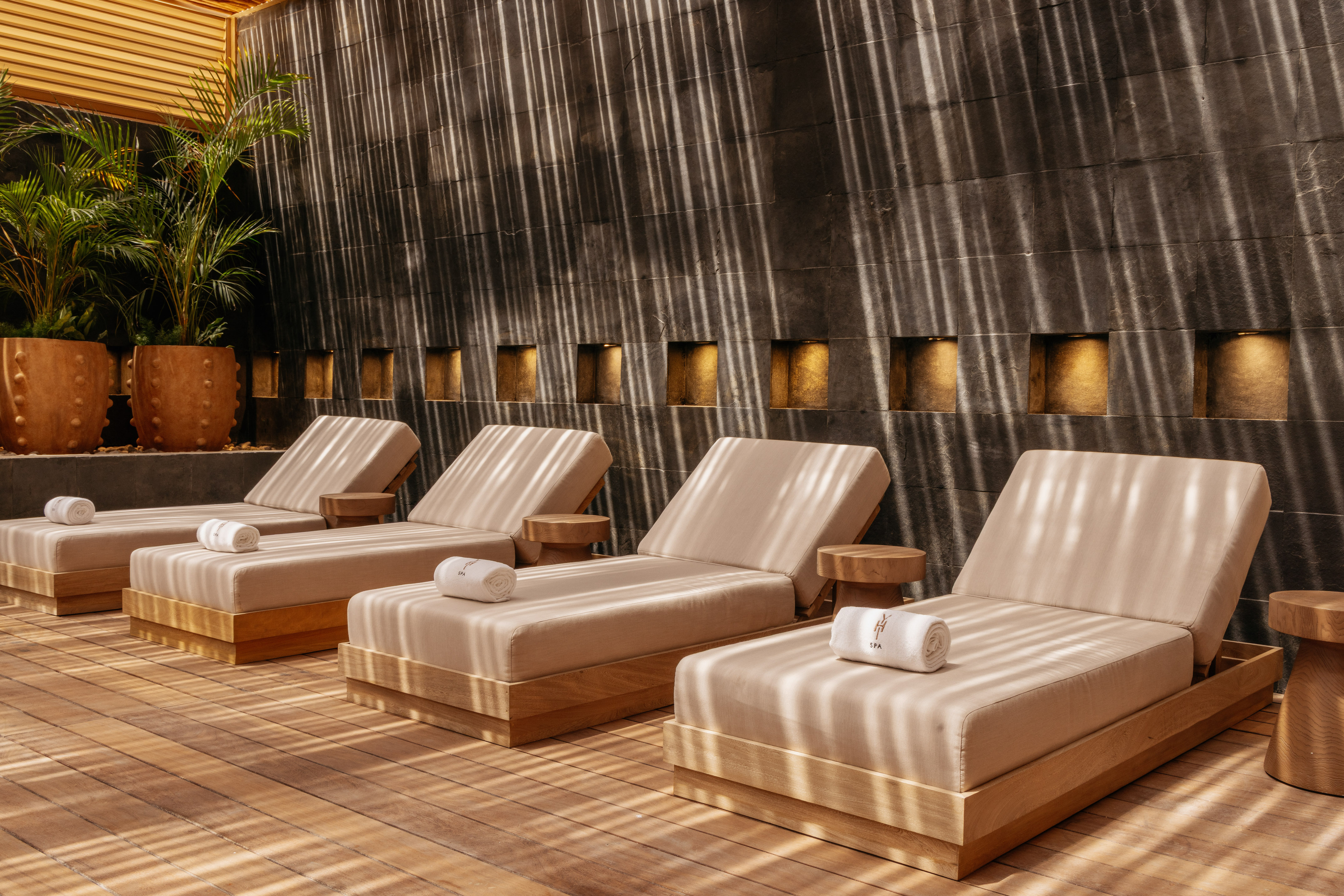 Spa daybeds on wooden deck, dark stone wall, palm plants, sun stripes.