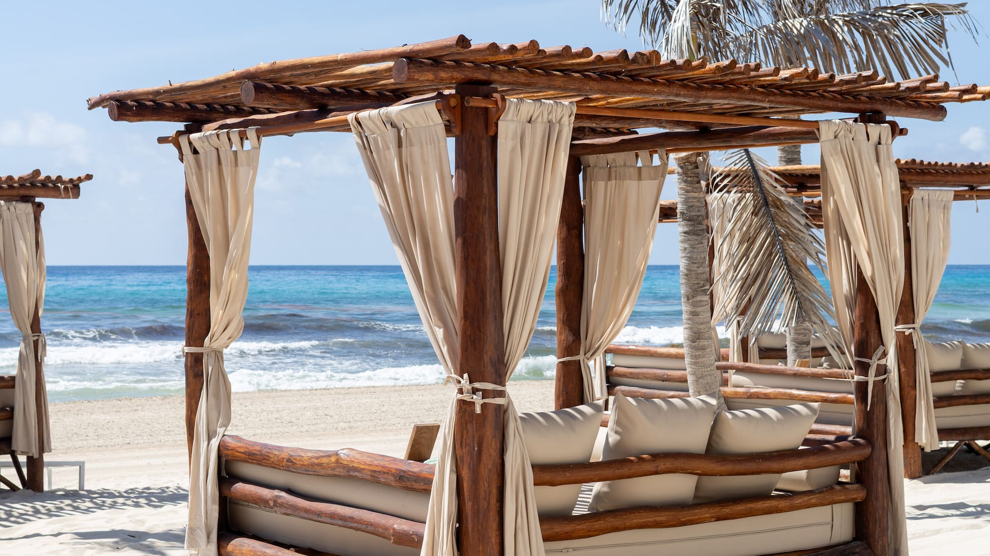 Wooden beach cabana, white daybed on sand, blue ocean, palm trees.