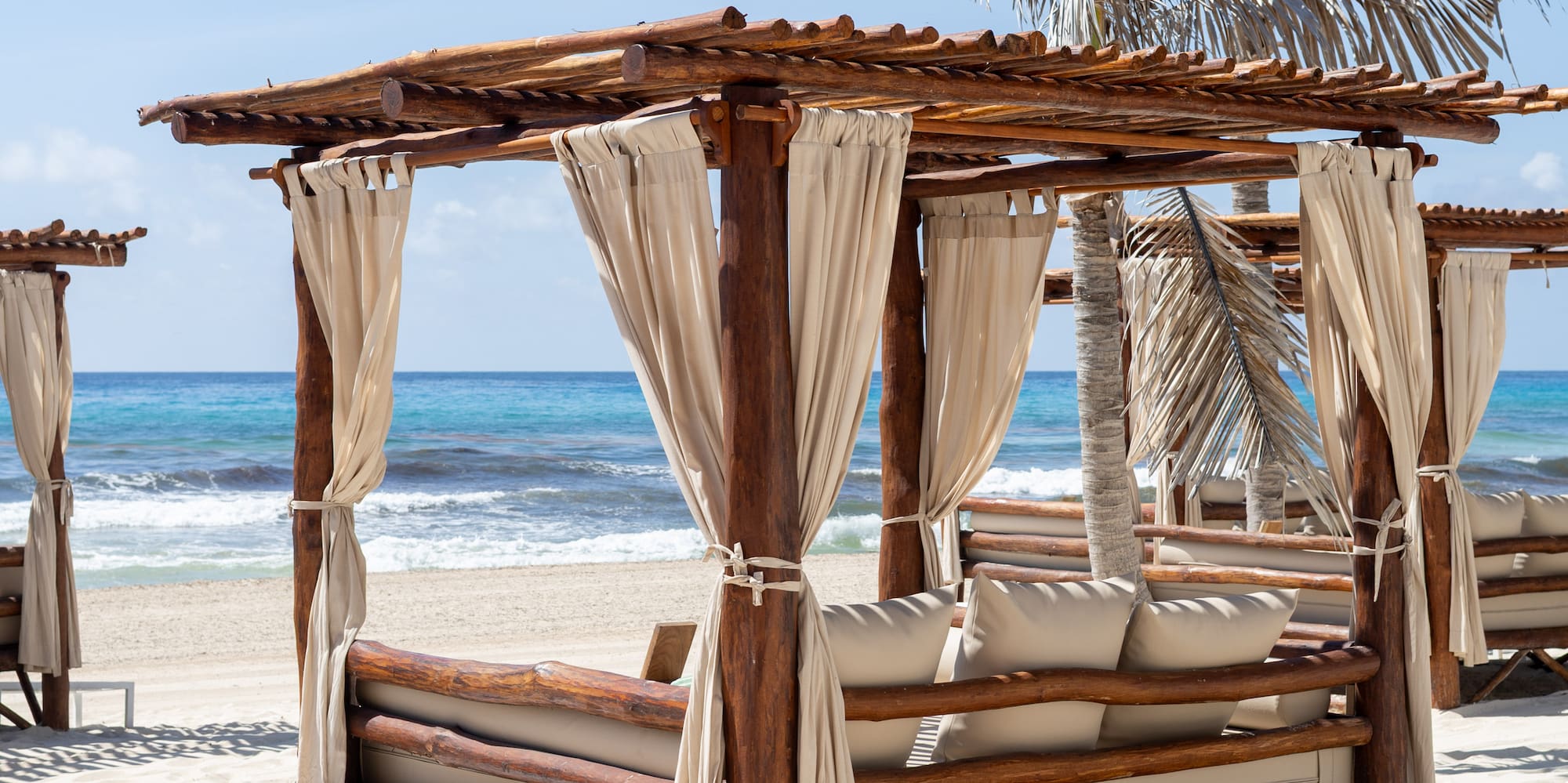 Wooden beach cabana, white daybed on sand, blue ocean, palm trees.