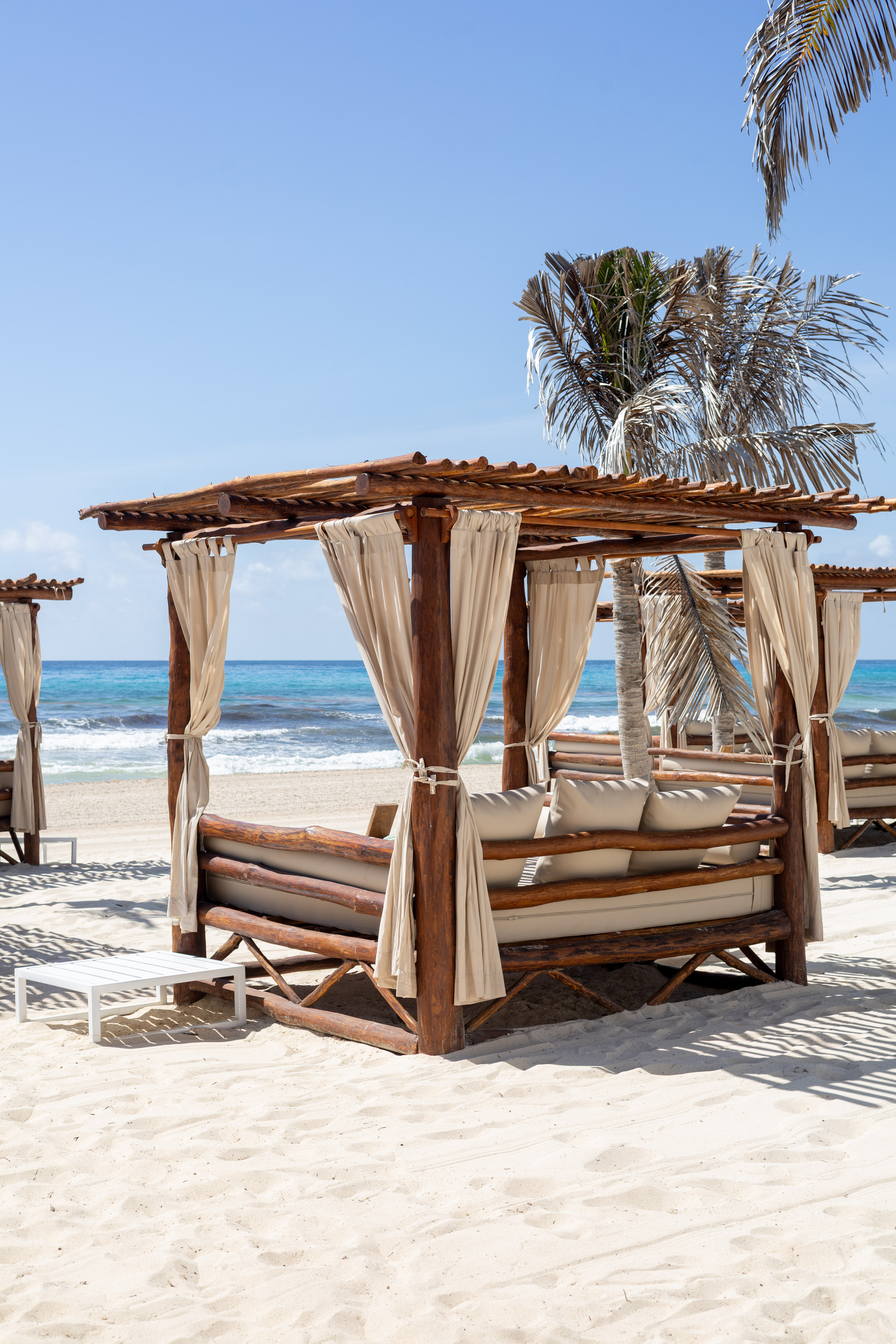 Wooden beach cabana, white daybed on sand, blue ocean, palm trees.