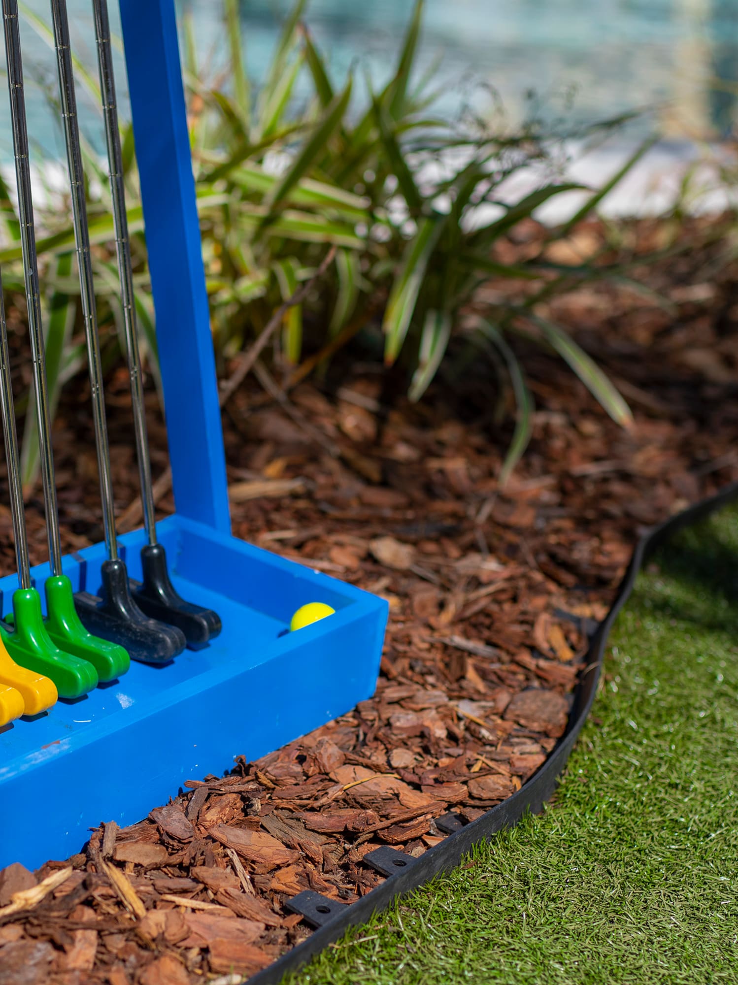 Colorful mini golf clubs in blue stand, yellow ball on artificial putting green.